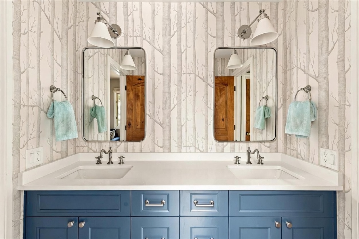 This double-vanity bathroom features a striking blue cabinet base topped with a white countertop and dual undermount sinks. The walls are adorned with a whimsical tree-patterned wallpaper, complemented by two rectangular mirrors and matching wall-mounted light fixtures. The space is finished with polished metal hardware and light blue hand towels, creating a clean and cohesive aesthetic.