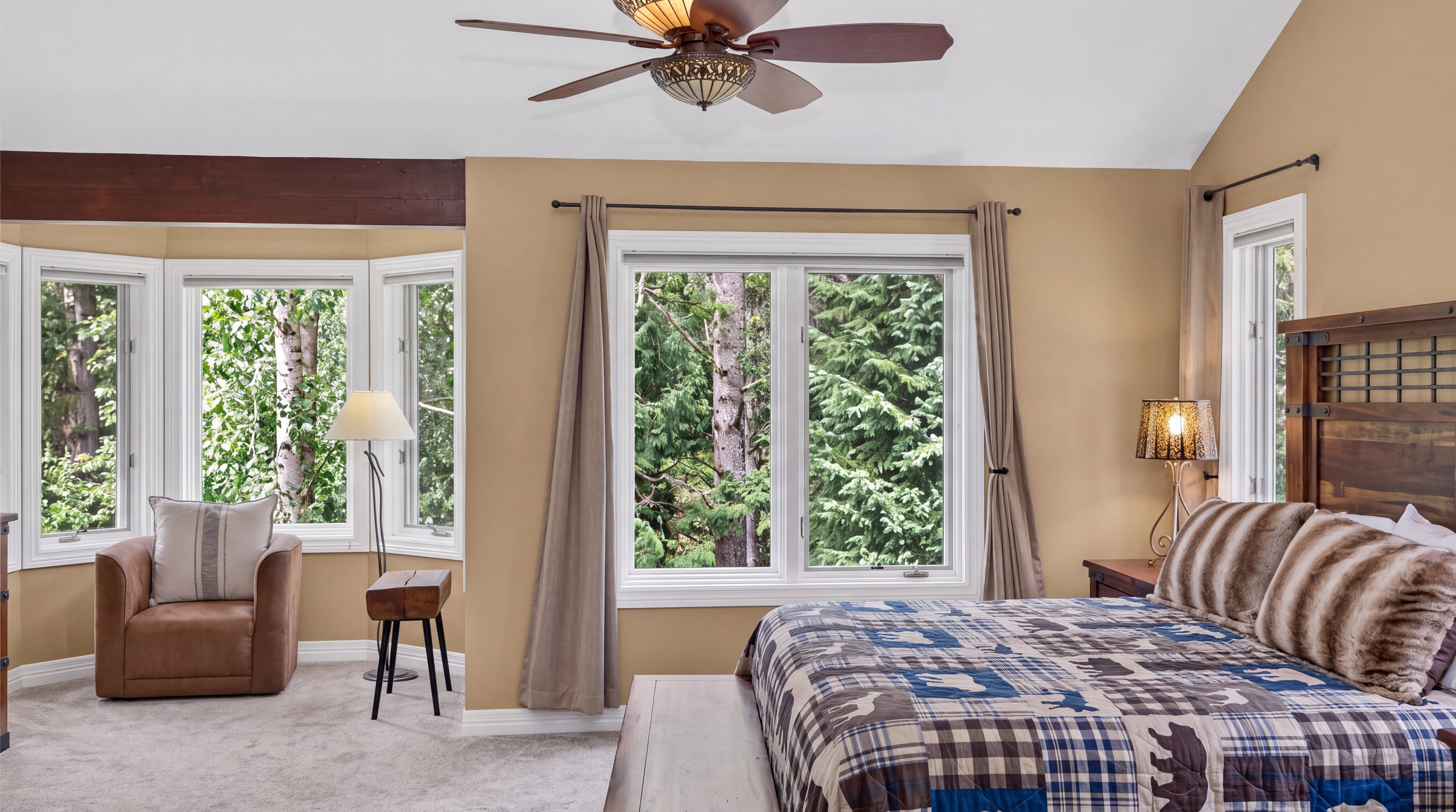 This cozy primary bedroom features a rustic design aesthetic with warm-toned walls, a vaulted ceiling, and an exposed wooden beam. The space is furnished with a comfortable bed adorned in a wildlife-themed quilt, a brown armchair, and a small side table, all overlooking scenic forest views through expansive windows. The overall ambiance is inviting and tranquil, perfectly suited for a mountain or woodland retreat.