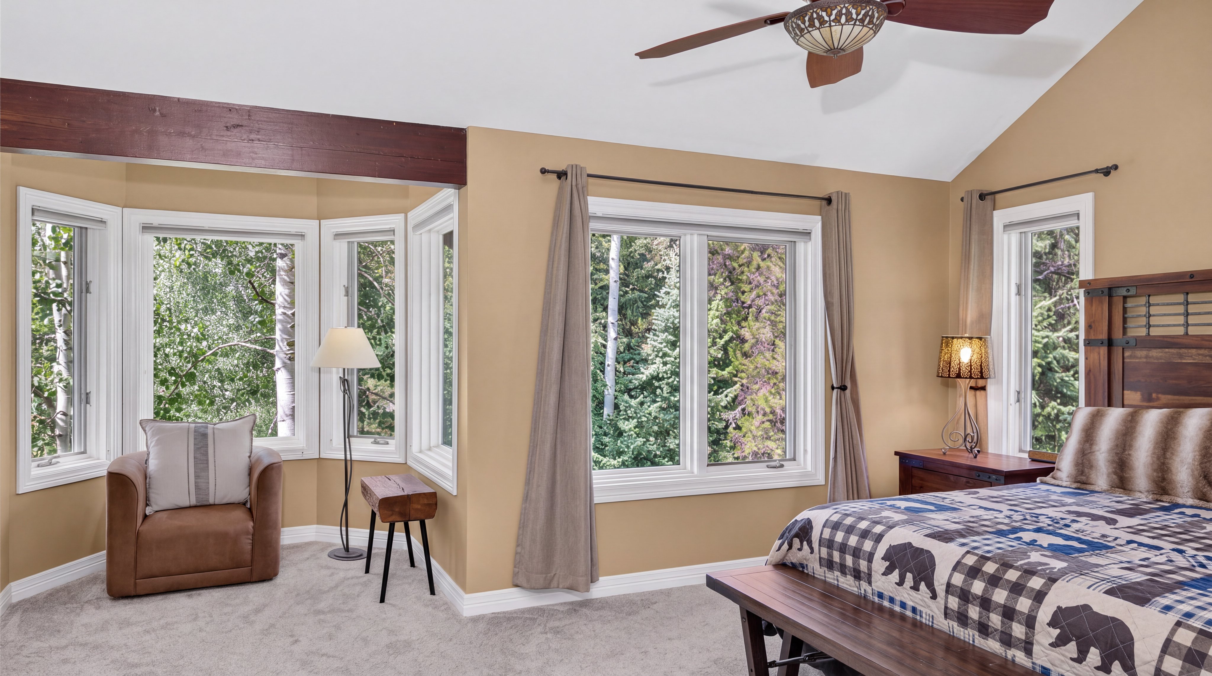 This spacious primary bedroom features a rustic-inspired decor with warm, earthy tones and an inviting atmosphere. The room is highlighted by a cozy reading corner with a leather armchair, a beautiful quilt with bear motifs, and multiple windows that provide ample natural light and scenic woodland views. High ceilings with a wooden beam and a central ceiling fan add to the charming, cabin-like aesthetic of the space.