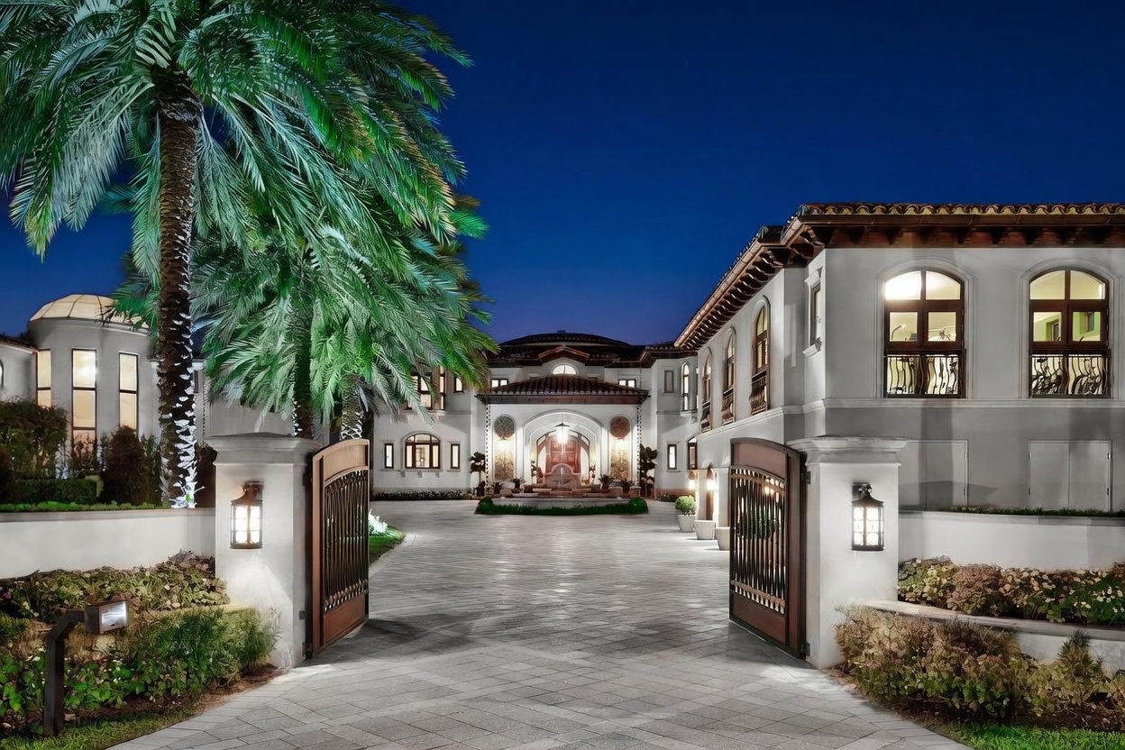 This grand Mediterranean-style estate is captured at dusk, showcasing an impressive gated entrance with a paved driveway leading to a central fountain. The symmetrical architecture features arched windows, warm exterior lighting, and lush landscaping, creating a sophisticated and welcoming curb appeal. The perspective is a wide, eye-level shot that emphasizes the scale and luxury of the property's facade.
