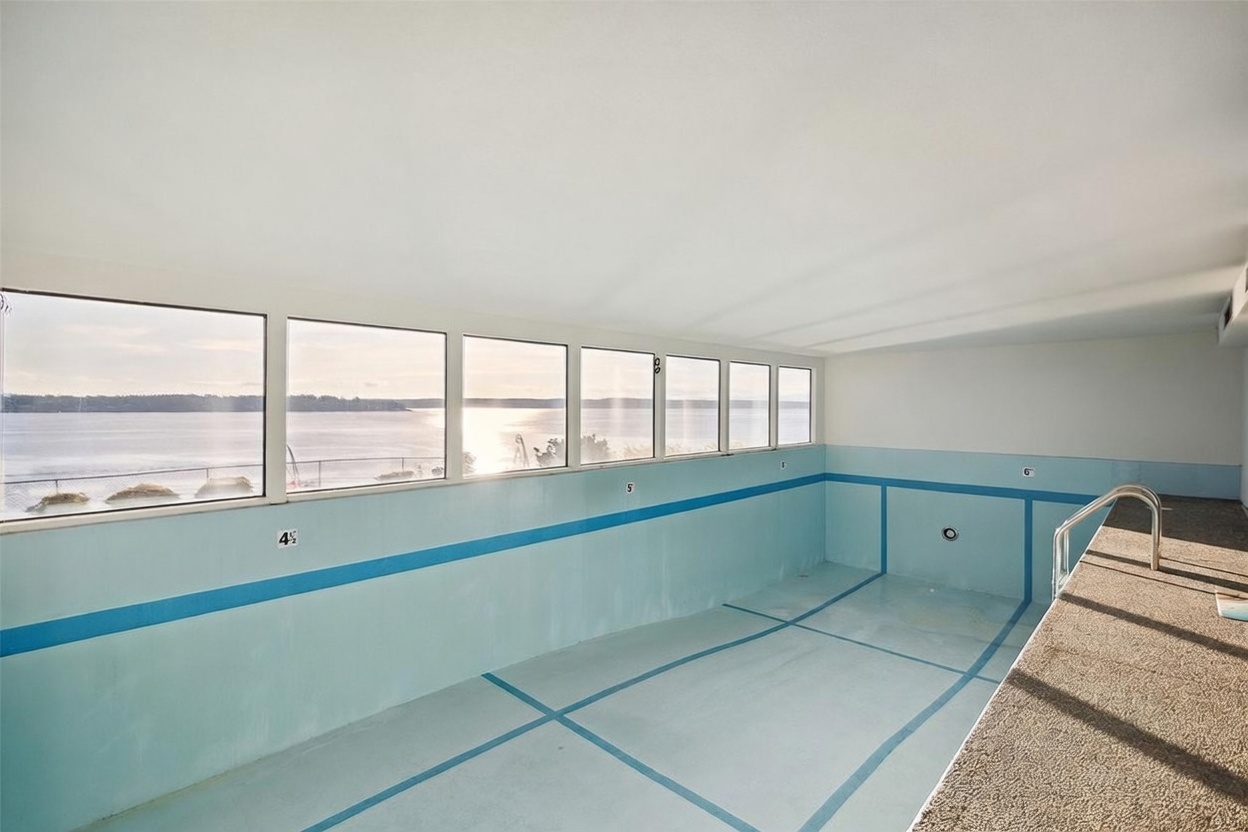 This indoor pool area features a unique, elongated design with depth markers painted along the light blue walls. A series of large windows lines the exterior wall, providing an expansive, unobstructed view of the water and horizon outside. The pool deck is finished with a textured, speckled material, and a metal ladder provides entry into the water.