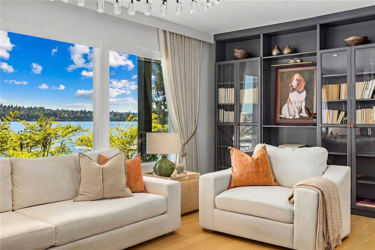 This elegant living room features a neutral color palette with plush seating and a large, dark-toned built-in shelving unit that displays decorative items and a portrait of a dog. Large windows provide an expansive view of the lake and surrounding greenery, filling the space with natural light. A crystal chandelier hangs above, adding a touch of sophistication to the comfortable and well-appointed seating area.