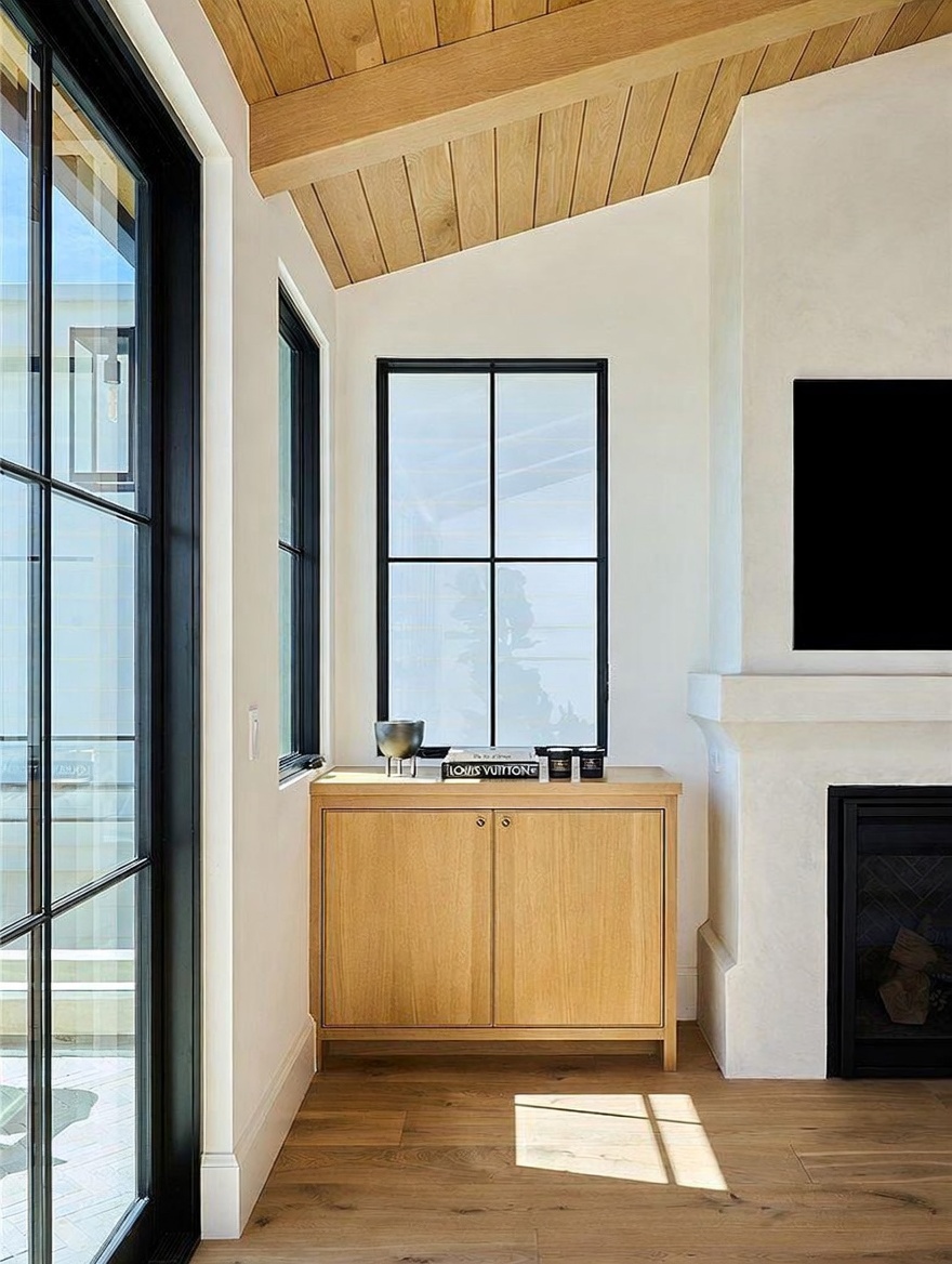 This interior space features a light wood cabinet positioned between a large window and a fireplace with a tall, smooth surround. The room is accented by a vaulted ceiling with light wood paneling and matching light wood flooring, creating a cohesive and airy aesthetic. The perspective captures a clean, modern living area with a focus on the architectural details and natural light.