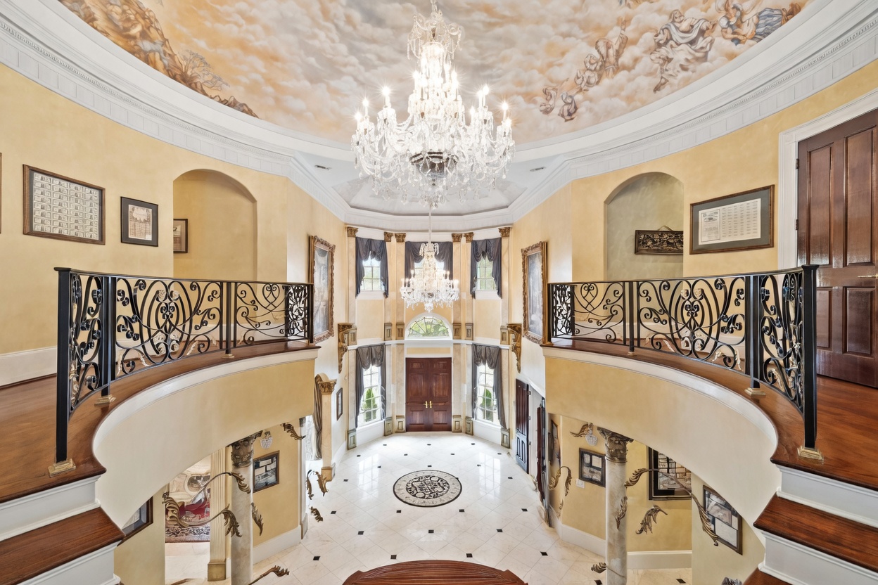 This grand foyer features a dramatic double staircase with intricate black wrought-iron railings and polished wood handrails. A massive crystal chandelier hangs from a ceiling adorned with a detailed mural, while the entryway below showcases a patterned tile floor and a set of double doors. The perspective is from the top of the stairs, looking down into the expansive, light-filled entrance hall.