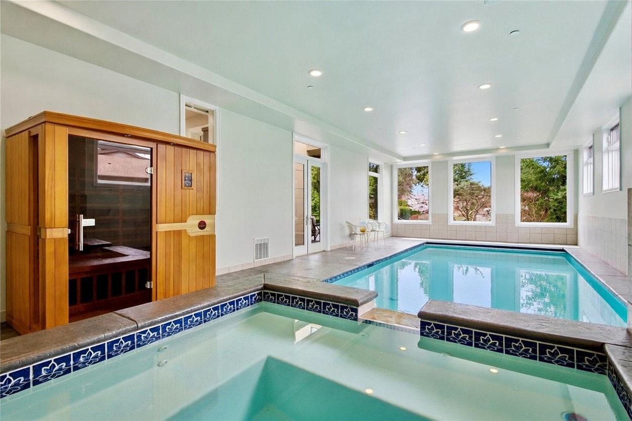 This indoor pool and spa room features a large, light-filled space with a built-in sauna and a tiled hot tub in the foreground. The room is surrounded by large windows that provide views of the outdoors, while recessed lighting illuminates the neutral-toned walls and tiled flooring. The overall atmosphere is one of a private, resort-style wellness retreat.