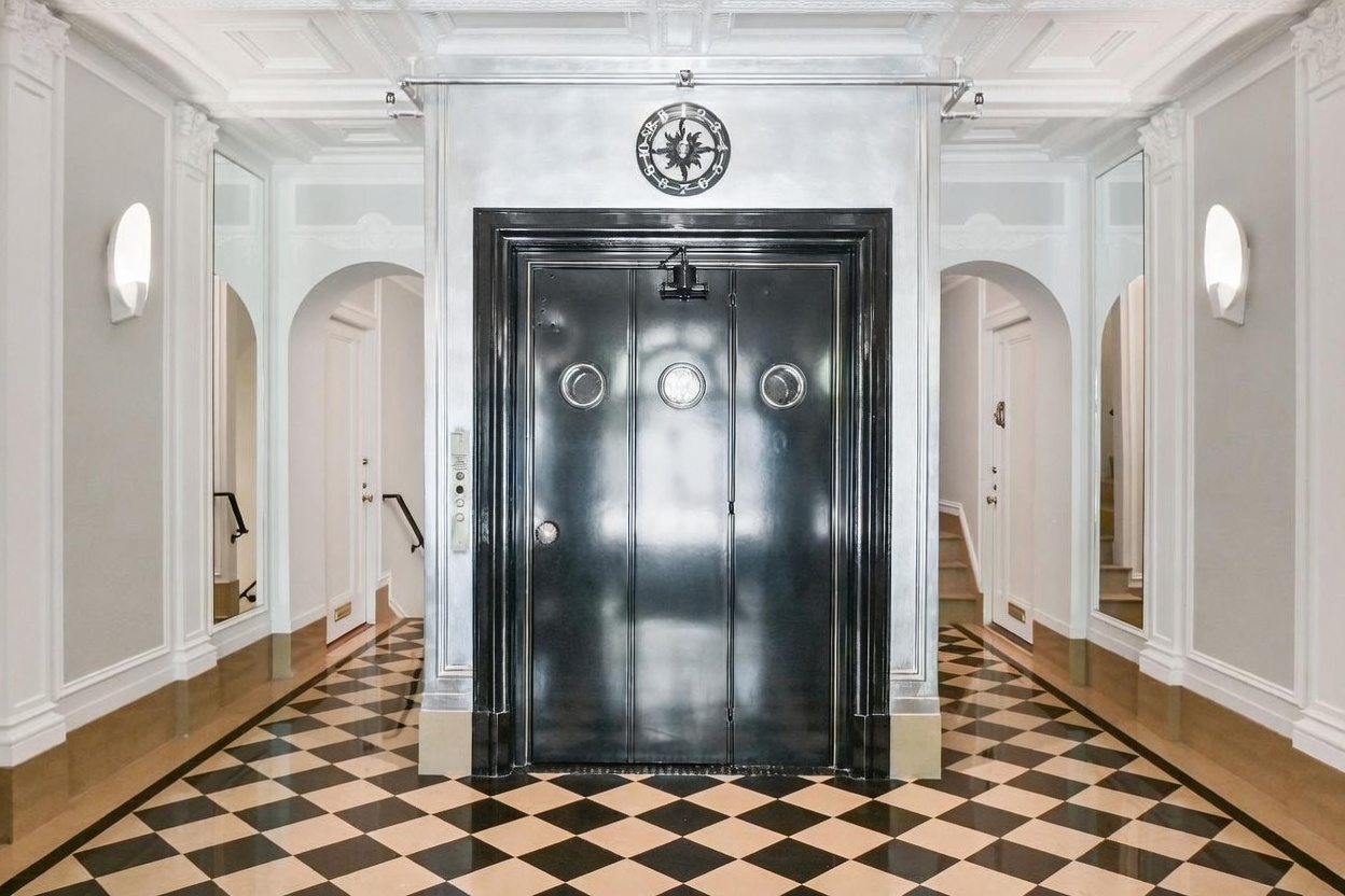 This elegant foyer features a striking black elevator door with circular windows, centered against a metallic-finished wall. The space is defined by a classic black-and-white checkered floor, symmetrical arched doorways leading to staircases, and ornate ceiling molding. Two wall-mounted sconces provide soft lighting, creating a sophisticated and dramatic entryway.