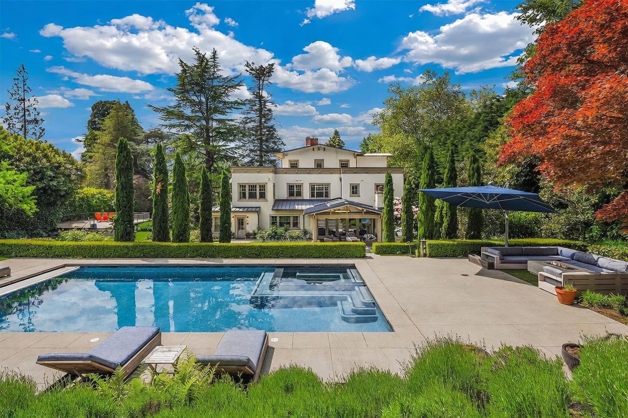 This expansive backyard features a large, rectangular swimming pool with an integrated spa, set against a backdrop of a multi-story, light-colored residence. The pool deck is paved with neutral stone, complemented by a comfortable outdoor lounge area shaded by a large blue umbrella and a manicured hedge line. The scene is framed by mature trees and a vibrant red-leafed tree, creating a serene and luxurious outdoor living environment.