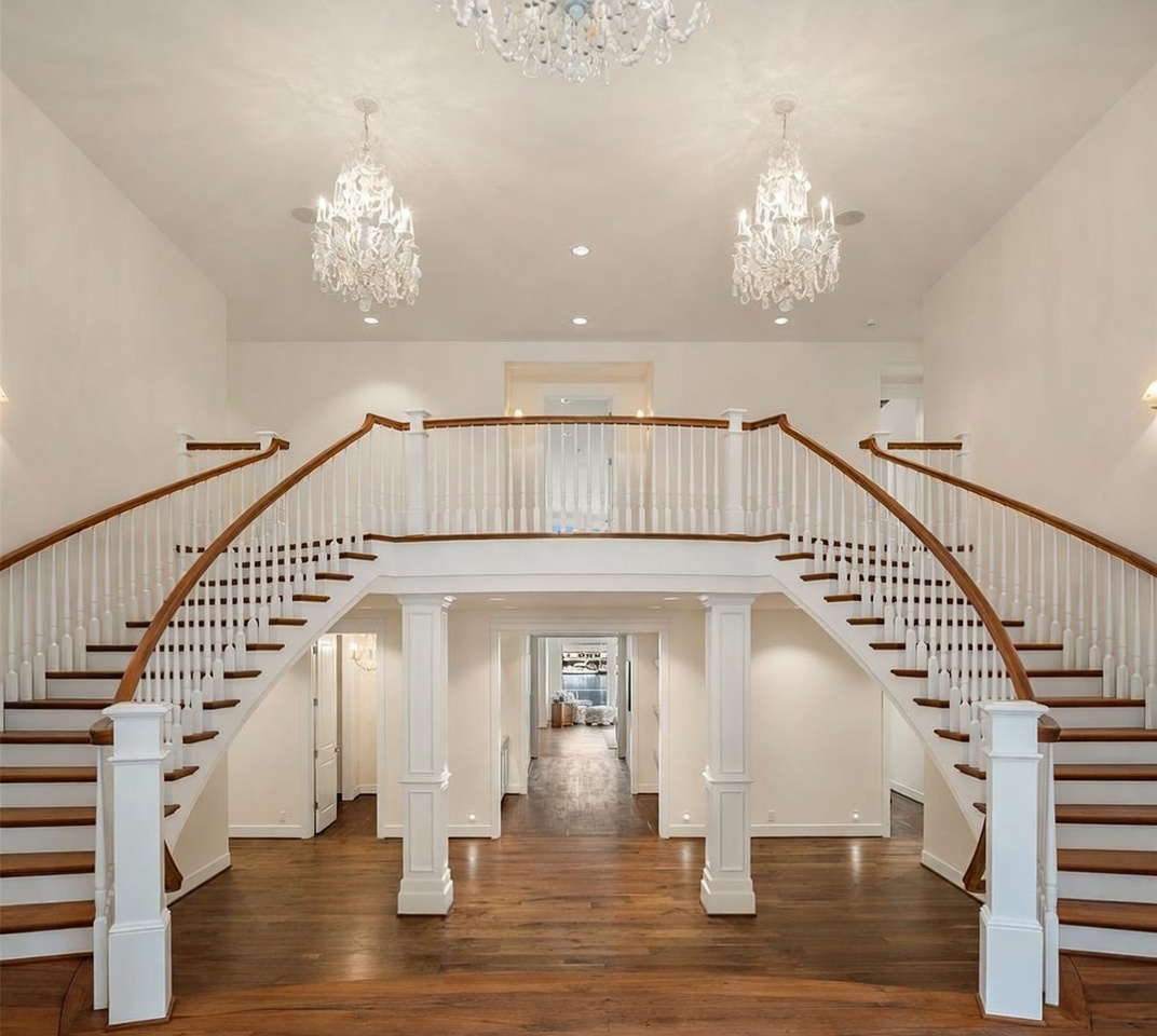 This grand foyer features a dramatic dual staircase with white balusters and wood handrails that curve upward to a central landing. The space is illuminated by three ornate crystal chandeliers suspended from the high ceiling, casting a warm glow over the dark wood flooring. Two prominent white columns frame the entrance to a central hallway, creating a symmetrical and elegant architectural focal point.