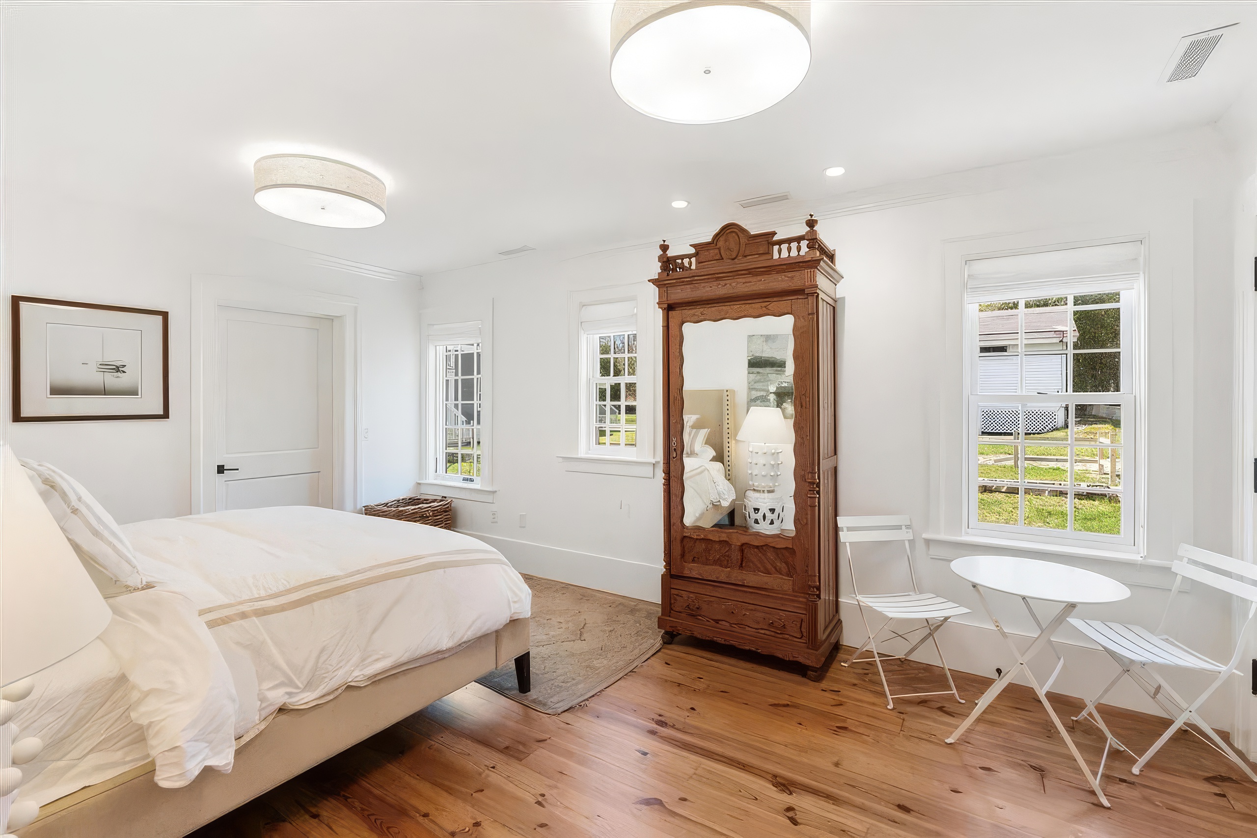 This bright and airy bedroom features light wood flooring and crisp white walls, creating a clean, minimalist aesthetic. The room is furnished with a comfortable bed, a large antique-style wooden armoire with a mirrored door, and a small bistro-style table with two chairs near the window. Large windows allow for ample natural light, providing a pleasant view of the outdoors.