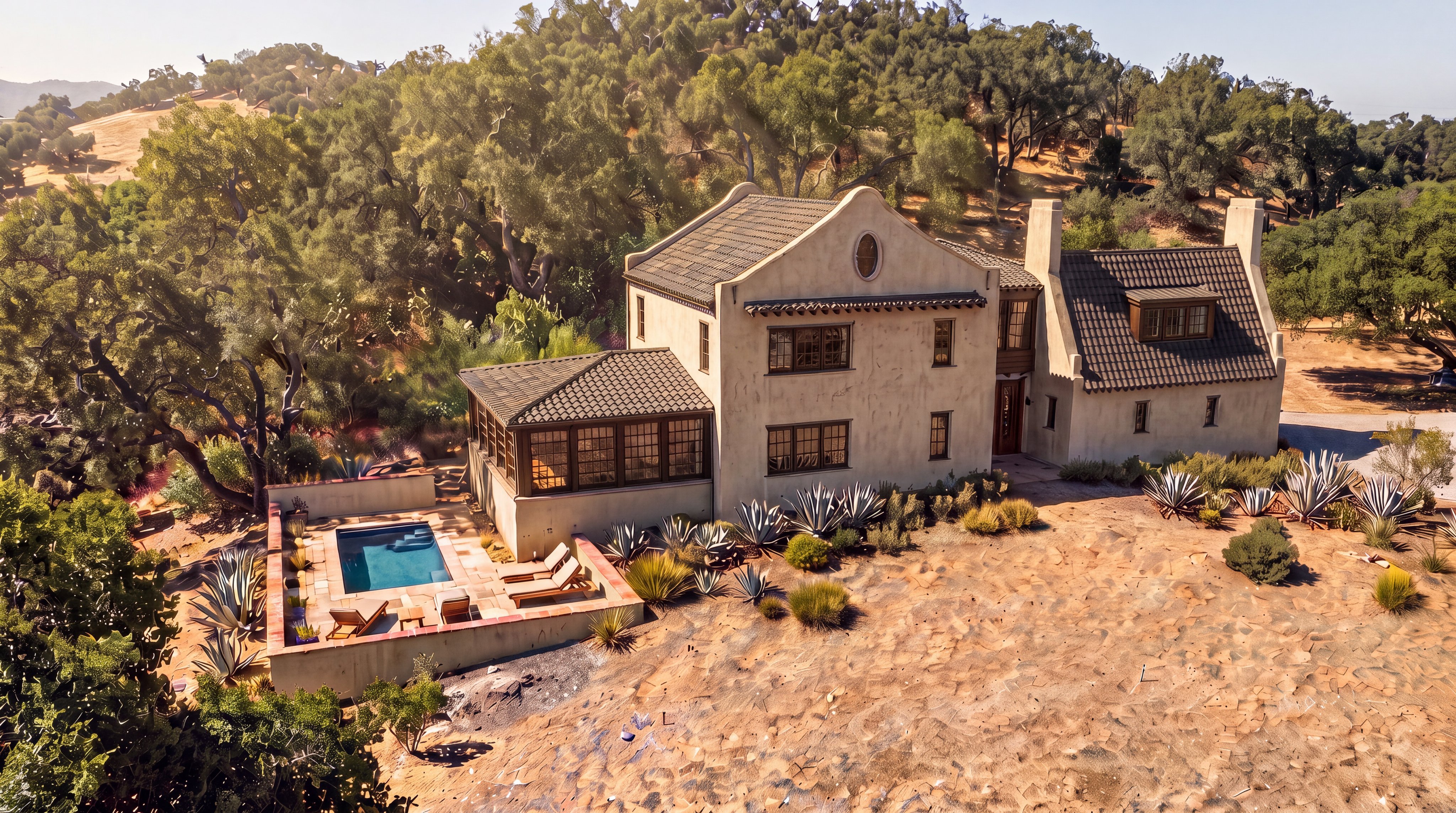 This stunning Mediterranean-style estate is captured from an elevated aerial perspective, showcasing its harmonious integration with the surrounding natural landscape. The home features a classic stucco finish, dark tiled roofing, and a charming sunroom addition, complemented by a serene private pool area with lounge seating. The surrounding desert foliage and gentle hillside backdrop create an inviting and tranquil atmosphere, characteristic of high-end, secluded properties.