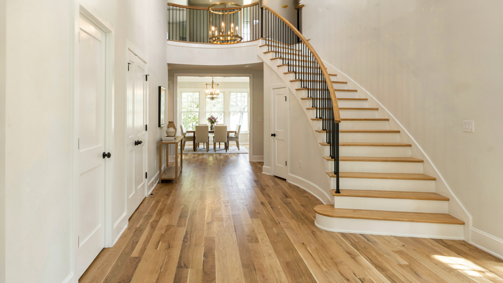 This grand foyer showcases a dramatic curved staircase with wooden treads and black iron railings, serving as the focal point of the entryway. The space features elegant hardwood flooring throughout and leads directly into a bright dining area, creating an inviting and open-concept flow. Neutral wall tones and classic millwork enhance the home's sophisticated, high-end aesthetic.