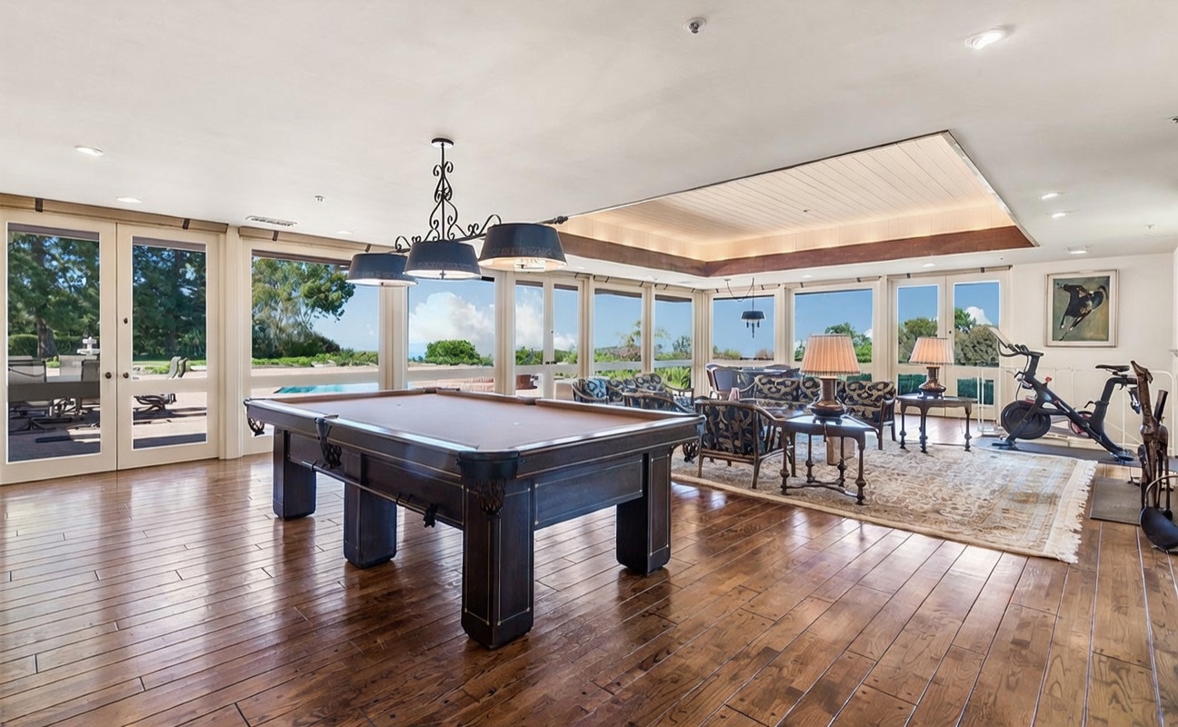 This expansive sunroom features rich wood flooring and a large pool table as the central focal point. The space is surrounded by large windows that provide panoramic views of the outdoor landscape and pool area, creating a bright and airy atmosphere. A comfortable seating area with patterned furniture and a stationary exercise bike are arranged to take advantage of the natural light and open layout.