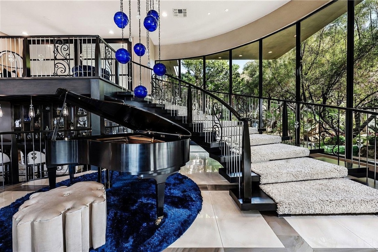 This dramatic living space features a grand piano positioned on a circular blue rug, set against a sweeping staircase with ornate metal railings. The room is defined by a curved wall of floor-to-ceiling windows that provide an expansive view of the lush, wooded exterior. A striking blue glass orb chandelier hangs from the ceiling, adding a modern artistic focal point to the sophisticated, open-concept interior.