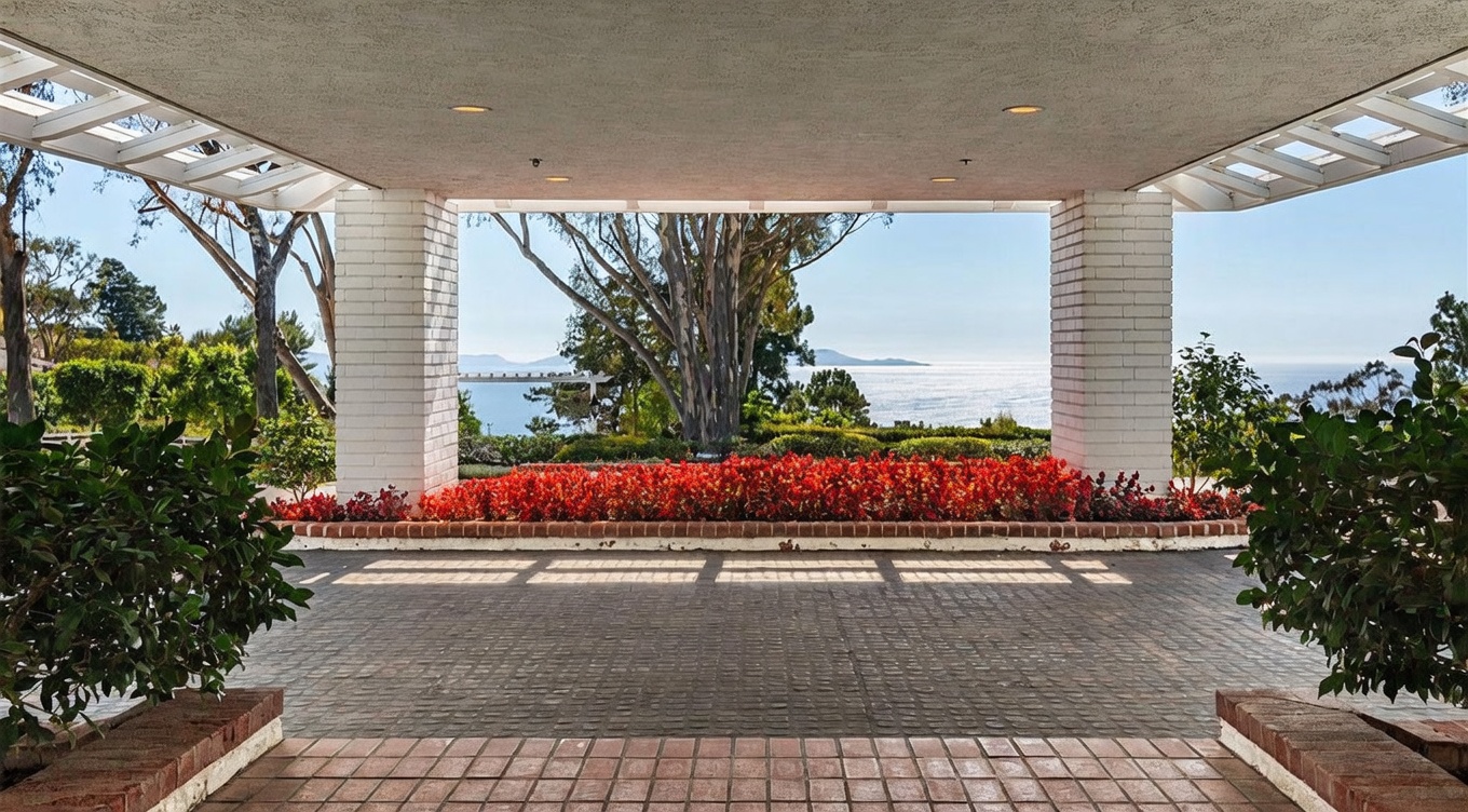 This grand porte-cochère serves as a dramatic entryway, featuring white brick pillars and a covered ceiling with recessed lighting. The space opens up to a stunning, unobstructed view of the ocean and distant coastline, framed by lush greenery and a vibrant bed of red flowers. The paved driveway leads directly toward the horizon, creating an impressive and welcoming arrival experience.