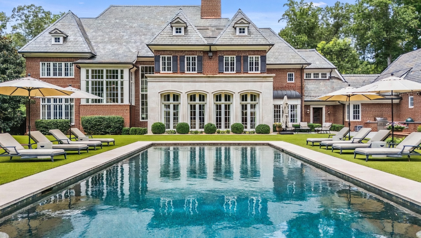 This rear view of a grand brick estate showcases a symmetrical architectural design with a slate roof and multiple dormer windows. A large, rectangular swimming pool serves as the focal point of the manicured lawn, flanked by rows of lounge chairs and patio umbrellas. The central section of the home features tall, arched windows and a light-colored stone facade that contrasts elegantly with the surrounding red brick.