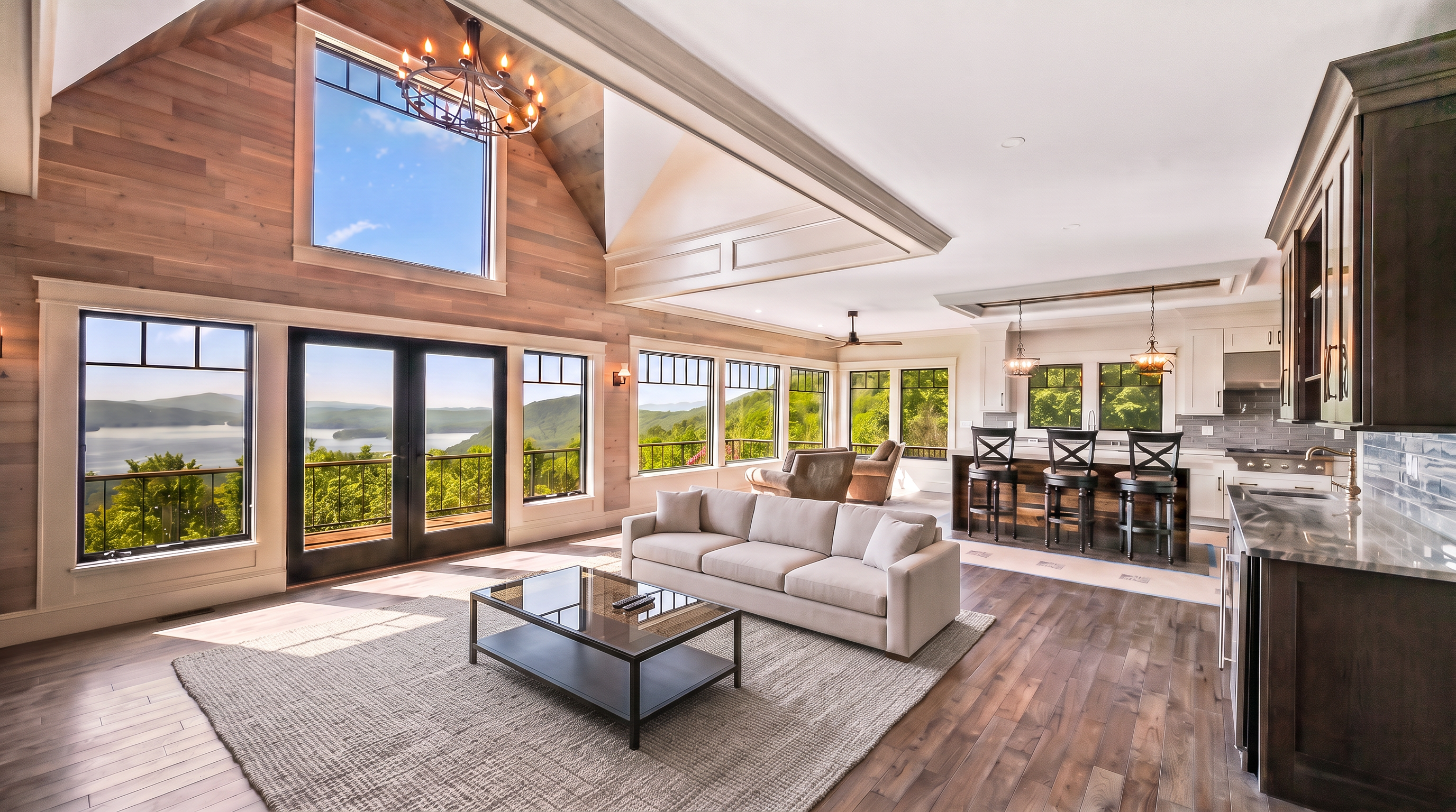 This spacious, open-concept living area features a grand vaulted ceiling with wood paneling and large floor-to-ceiling windows that capture an expansive mountain and lake view. The room is anchored by a contemporary sectional sofa, a coffee table, and a seamless flow into a gourmet kitchen with a breakfast bar. The warm, inviting aesthetic is enhanced by hardwood flooring and sophisticated lighting fixtures, making it perfect for both relaxation and entertaining.
