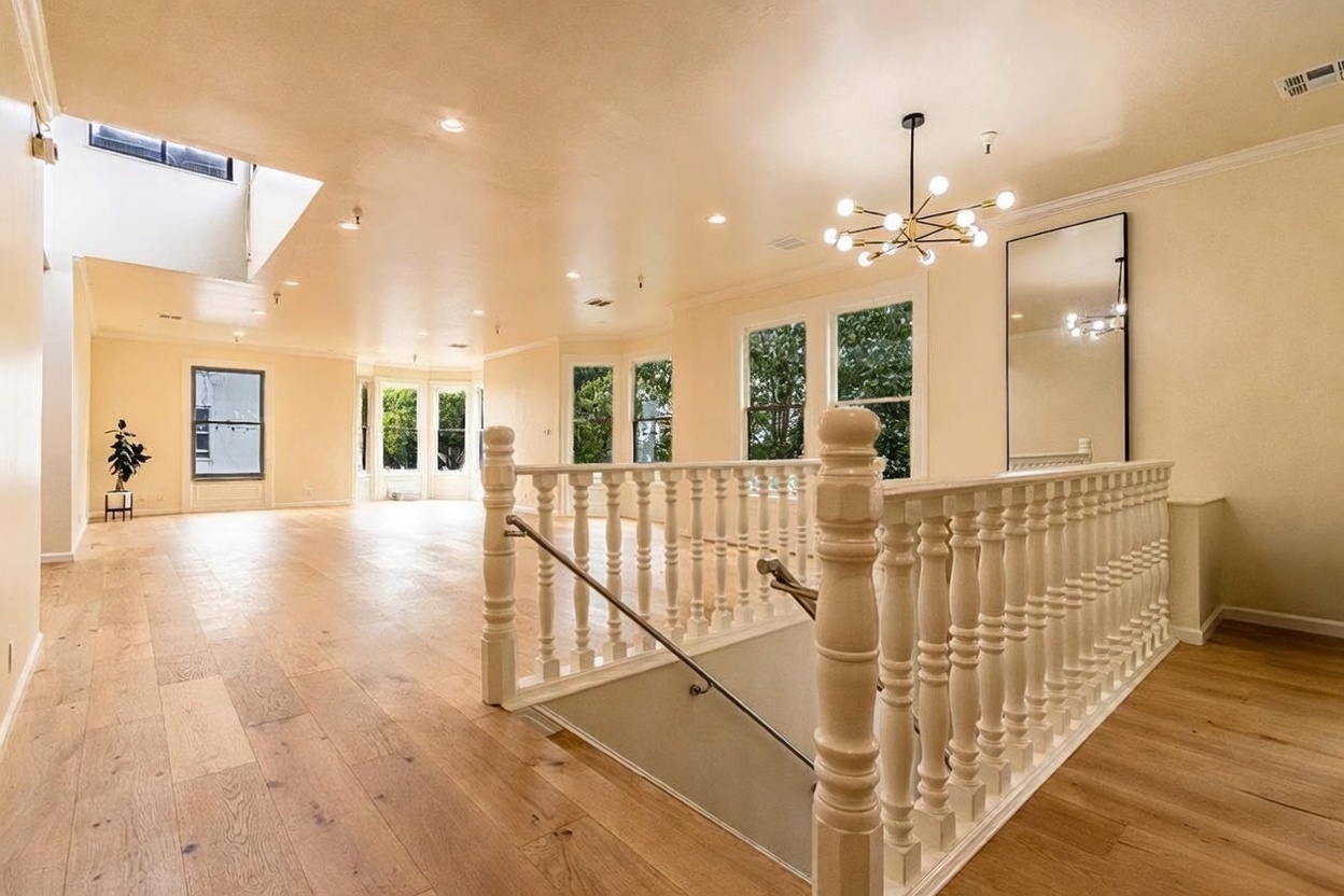 This bright, open-concept landing area features light wood flooring and a classic white balustrade overlooking the staircase. A modern, multi-bulb chandelier serves as a focal point, while large windows allow ample natural light to fill the space. The neutral color palette and clean lines create an airy, inviting atmosphere that connects the upper level to the rest of the home.