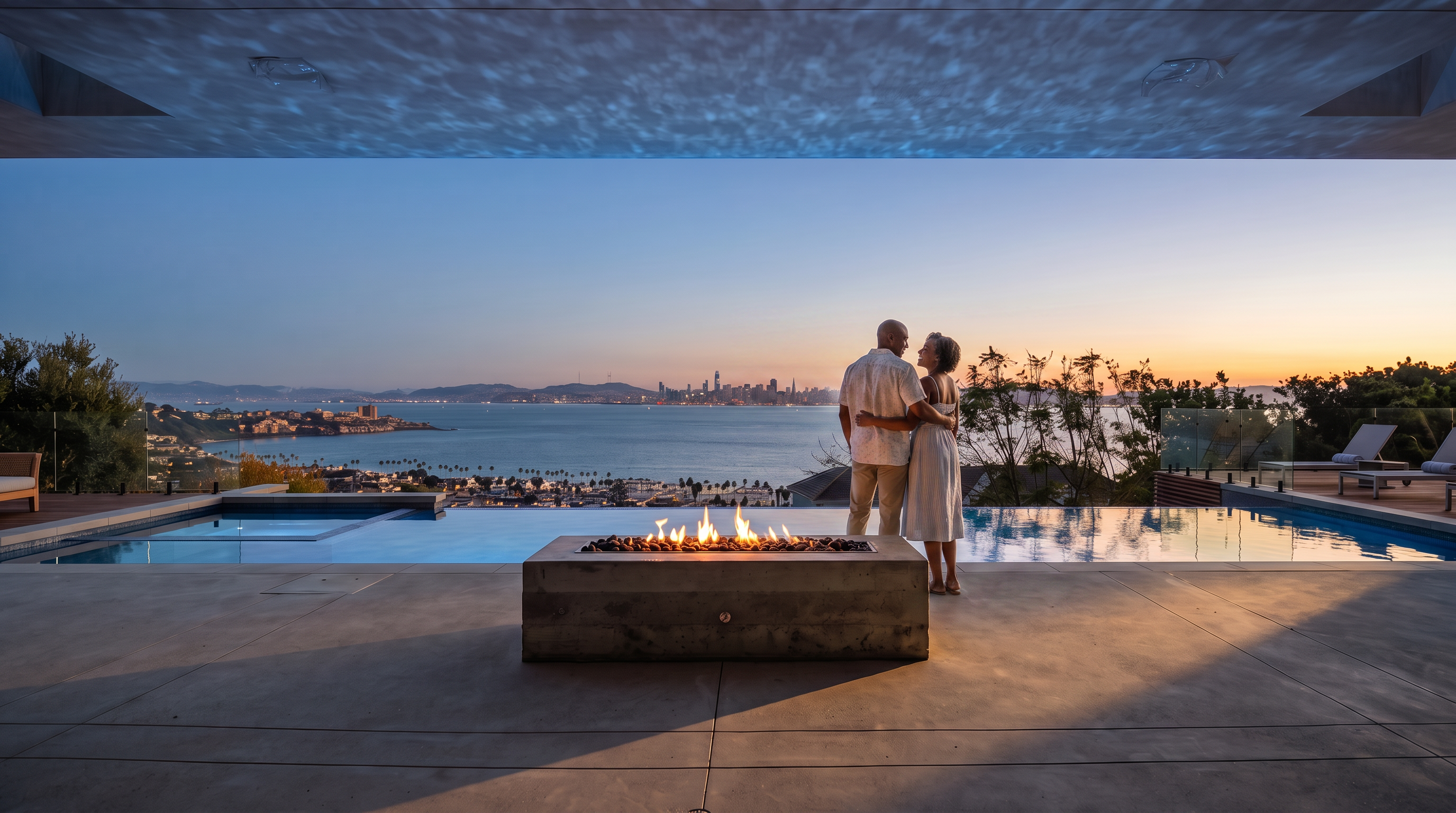 This stunning outdoor space features a sleek infinity-edge pool that seamlessly blends with the expansive ocean horizon in the background. A minimalist concrete fire pit sits on the foreground patio, providing a modern focal point for the terrace. The scene is framed by a covered patio area, offering a luxurious and serene atmosphere perfect for coastal living.