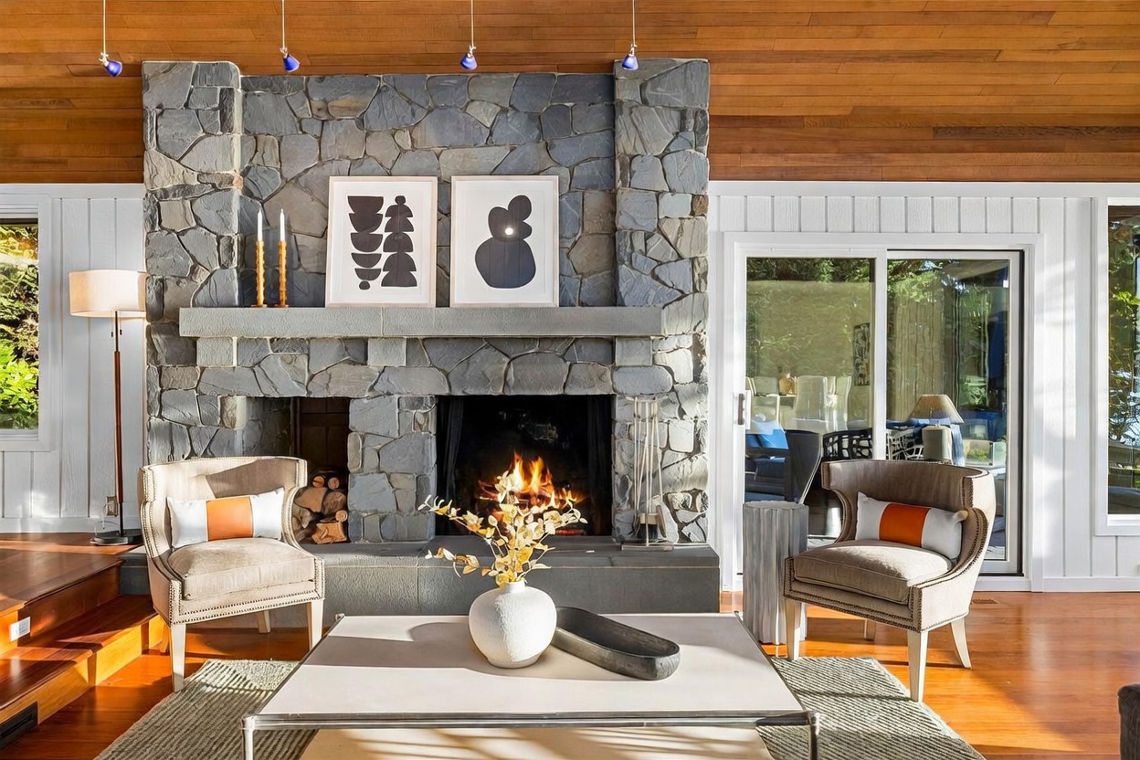 This inviting living room features a striking floor-to-ceiling stone fireplace as its centerpiece, flanked by two elegant armchairs with orange accent pillows. The space is warmed by a wood-paneled ceiling and rich hardwood floors, while large sliding glass doors provide a seamless connection to the outdoors. A modern coffee table with a decorative vase and a sleek tray completes the sophisticated, cozy atmosphere.