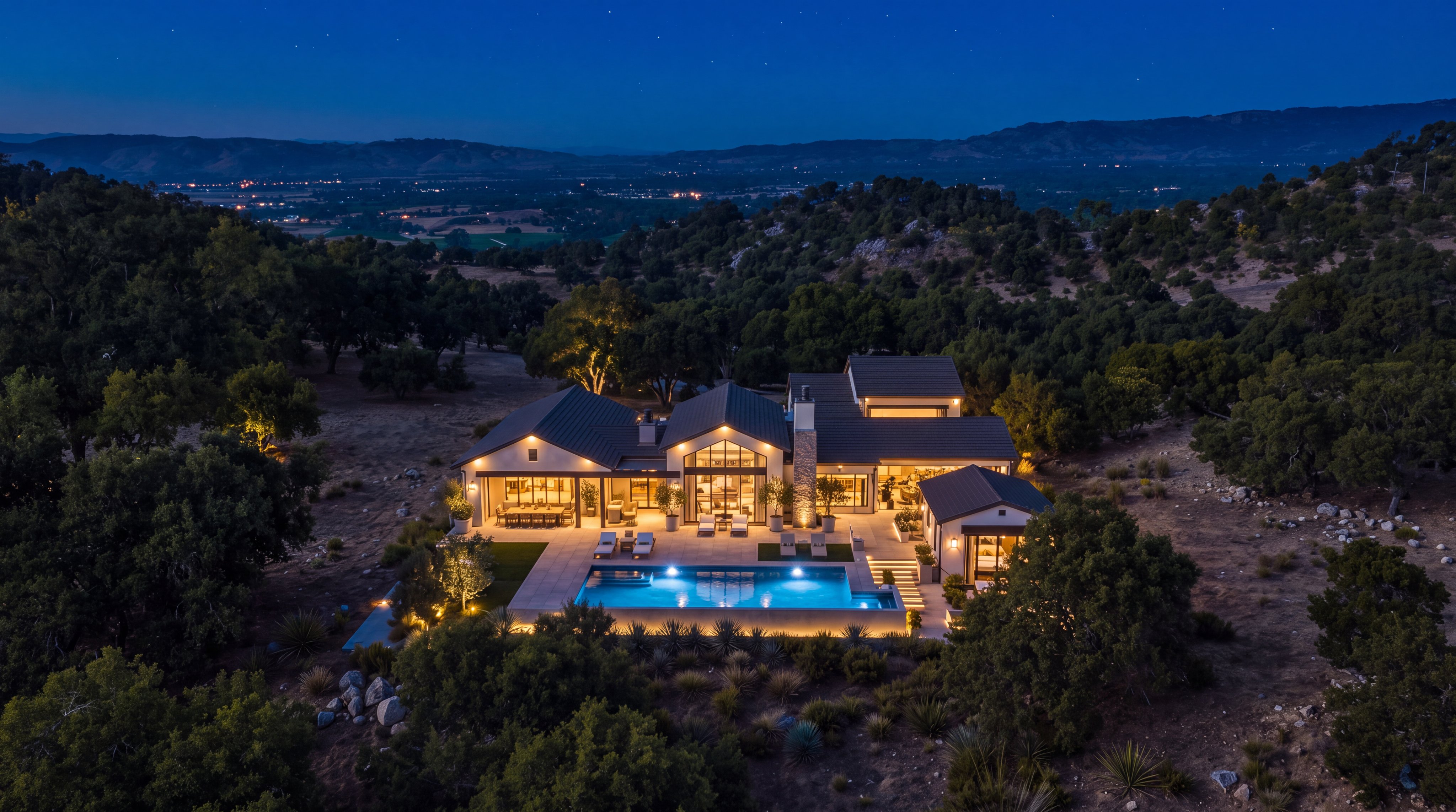 This striking aerial photograph captures a luxurious, modern estate at dusk, beautifully illuminated by interior and exterior lighting. The residence features a sophisticated architectural design with pitched roofs, expansive glass walls, and a prominent stone chimney, complemented by a pristine swimming pool and patio area. Nestled within a serene, wooded landscape with sweeping views of the distant valley, this property offers an exceptional blend of contemporary comfort and rural tranquility.