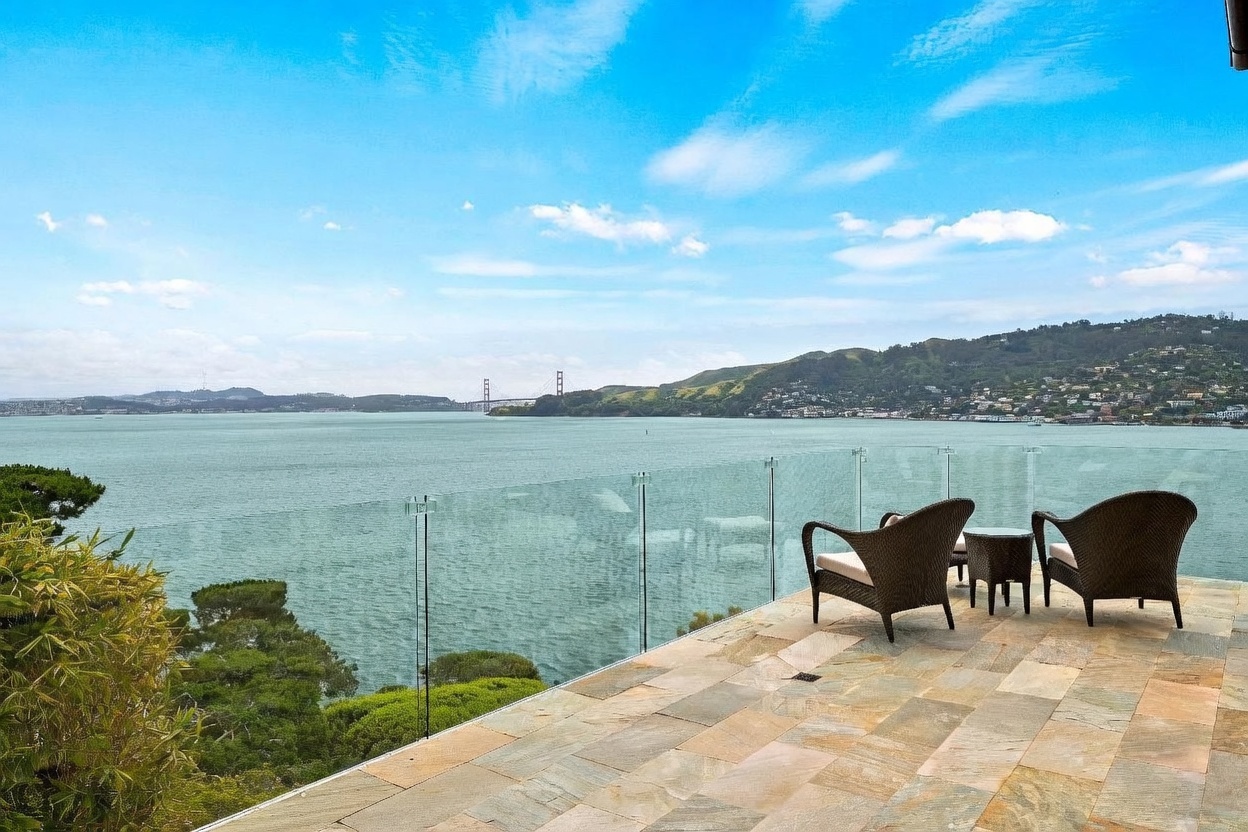 This expansive stone-paved terrace offers a breathtaking, unobstructed view of the bay and the distant Golden Gate Bridge. The space is furnished with two comfortable wicker armchairs and a small side table, all enclosed by a sleek, frameless glass railing that preserves the panoramic vista. The scene captures a serene, high-end outdoor living area perfectly positioned for enjoying the coastal scenery.