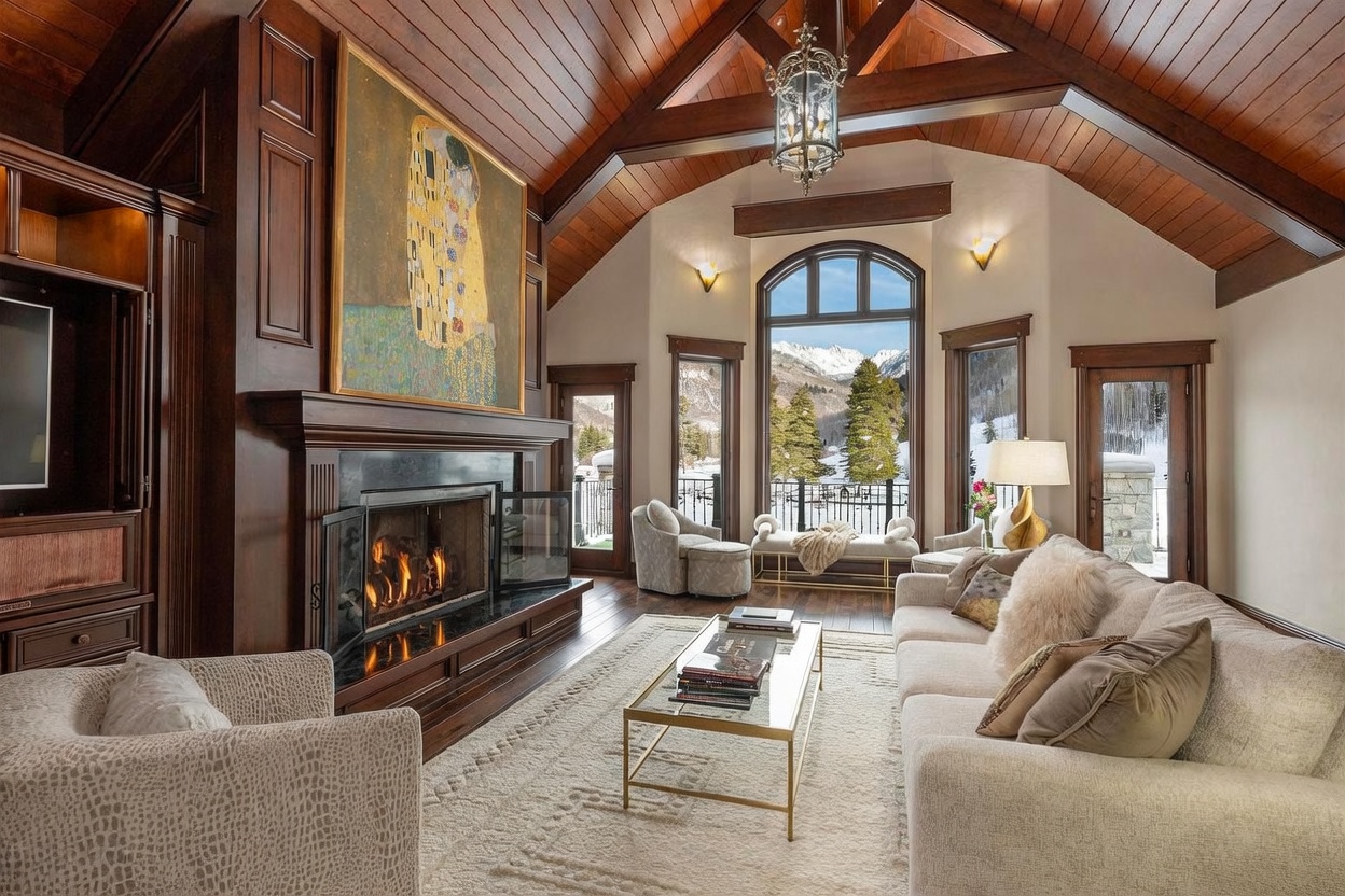 This elegant living room features a vaulted wood-paneled ceiling with exposed beams and a prominent fireplace with a dark stone surround and a large art piece above the mantel. A plush, light-colored sofa and matching armchairs are arranged on a textured area rug, creating a cozy seating area centered around a glass-topped coffee table. Large windows and glass doors provide expansive views of a snowy mountain landscape, filling the space with natural light and a sense of luxury.
