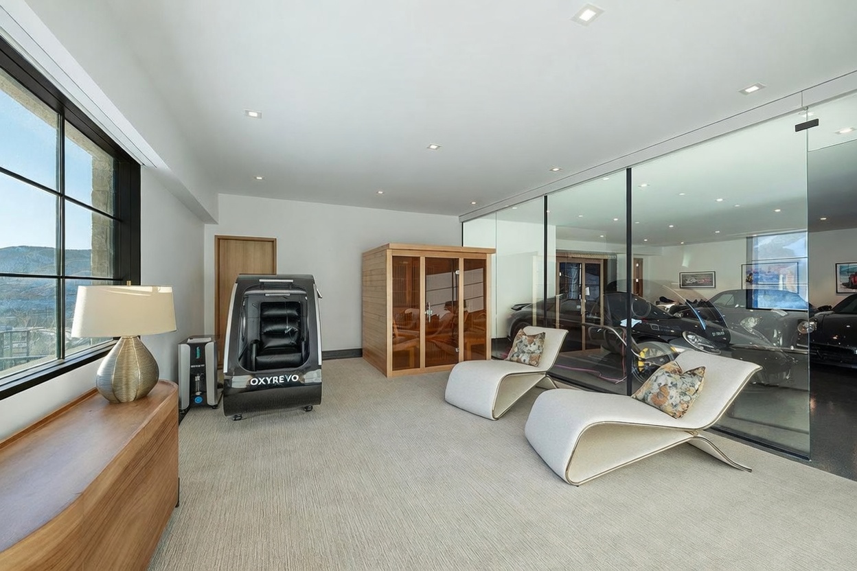 This modern home gym and wellness space features a sleek, minimalist aesthetic with light-toned carpeting and neutral walls. The room includes a hyperbaric oxygen chamber, a wooden sauna, and two contemporary lounge chairs, all positioned adjacent to a glass-walled garage display area showcasing luxury vehicles. Large windows on the left provide natural light and scenic views, creating a sophisticated and high-end atmosphere.