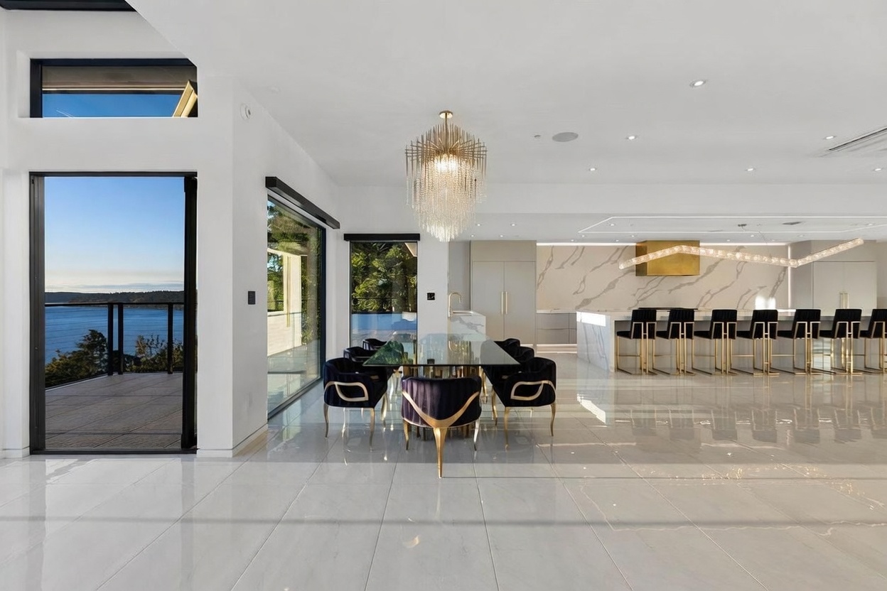 This open-concept dining area features a glass-topped table surrounded by dark velvet chairs, positioned beneath a dramatic tiered crystal chandelier. The space flows seamlessly into a sleek kitchen with a large island, gold-toned bar stools, and a striking stone-patterned backsplash. Large glass doors provide an expansive view of the water and deck, creating a bright, luxurious atmosphere with polished light-colored flooring throughout.