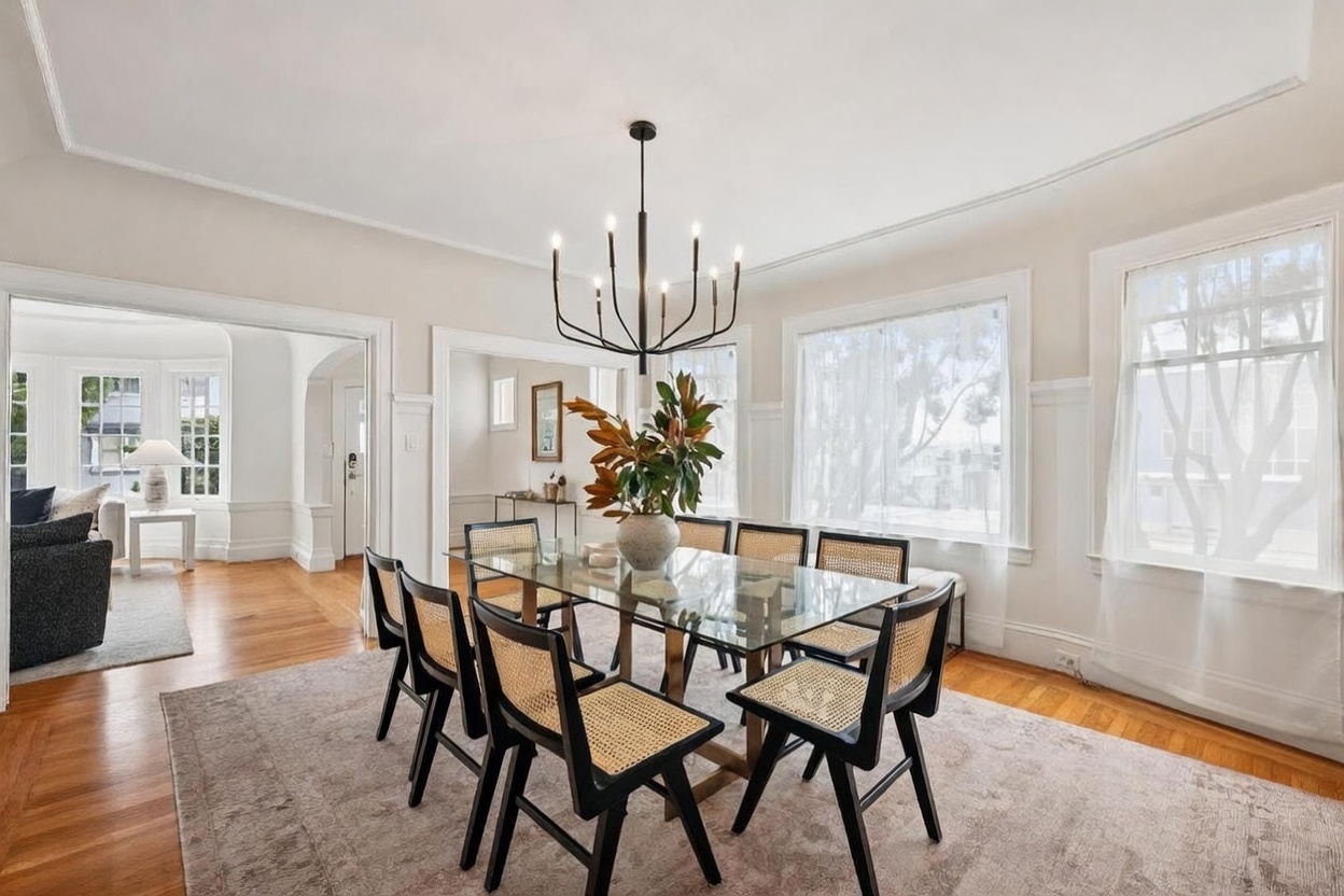 This elegant dining room features a glass-topped table surrounded by eight black chairs with woven cane seats, set atop a large, neutral-toned area rug. A modern, multi-armed black chandelier serves as a focal point above the table, while large windows with sheer curtains allow ample natural light to fill the space. The room showcases classic architectural details like crown molding and wainscoting, with a view into an adjacent living area through a wide doorway.