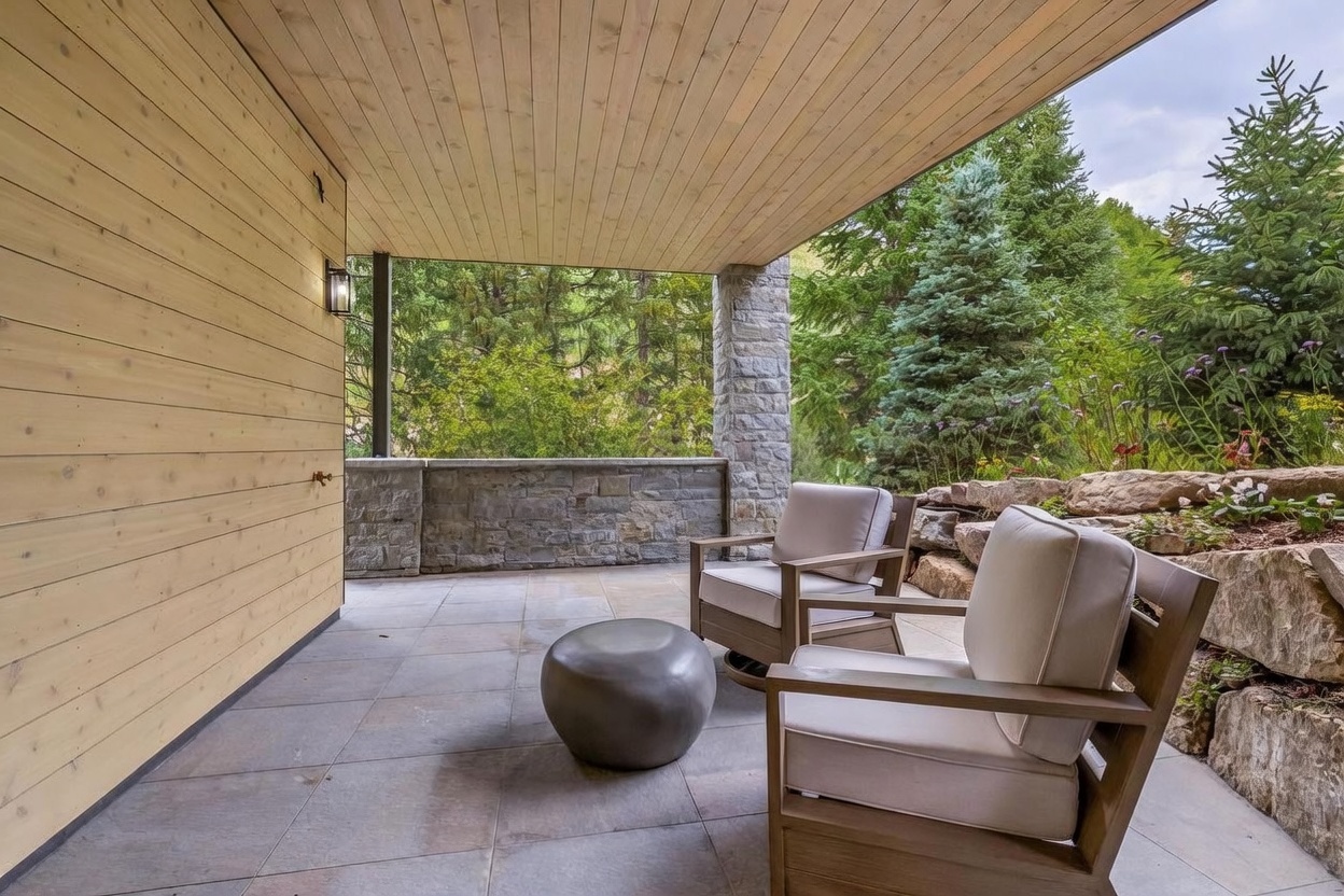 This covered patio features light wood-paneled walls and a stone-clad pillar, creating a warm and rustic aesthetic. Two comfortable armchairs with light-colored cushions are arranged around a low, rounded stone table, overlooking a lush, wooded landscape. The space is paved with large, neutral-toned tiles and offers a serene, private outdoor retreat.