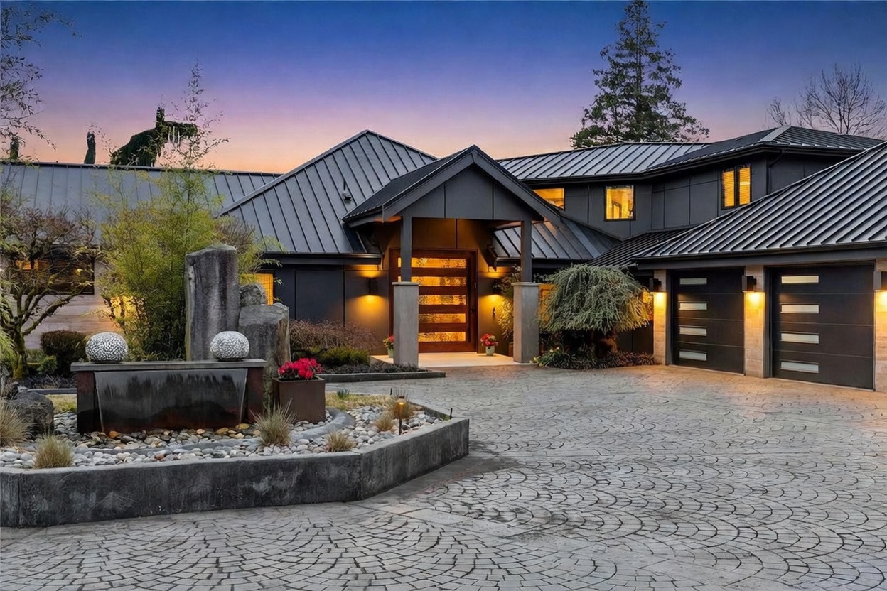 This striking modern residence features a dramatic dark metal roof and a sophisticated exterior with warm, inviting lighting that highlights the architectural lines. The front entrance is framed by a covered porch with stone pillars, leading to a grand wooden door, while a circular stone-paved driveway centers around a decorative water feature. The overall impression is one of luxury and contemporary elegance, captured at dusk to emphasize the home's curb appeal.