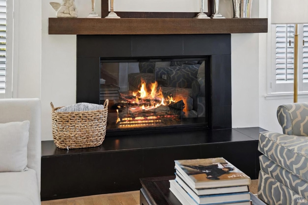 This cozy living room features a prominent fireplace with a dark, sleek surround and a substantial natural wood mantel. A woven basket sits on the hearth to the left, while a stack of books rests on a dark coffee table in the foreground. The room is framed by white walls and plantation shutters, creating a bright and inviting atmosphere.