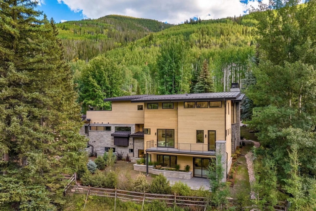 This elevated perspective captures a modern mountain residence nestled within a lush, forested landscape. The home features a blend of light wood siding and textured stone masonry, with a multi-level design that includes a spacious balcony and a ground-level patio. The surrounding dense trees and mountain backdrop create a serene, private atmosphere, highlighting the property's integration with its natural environment.