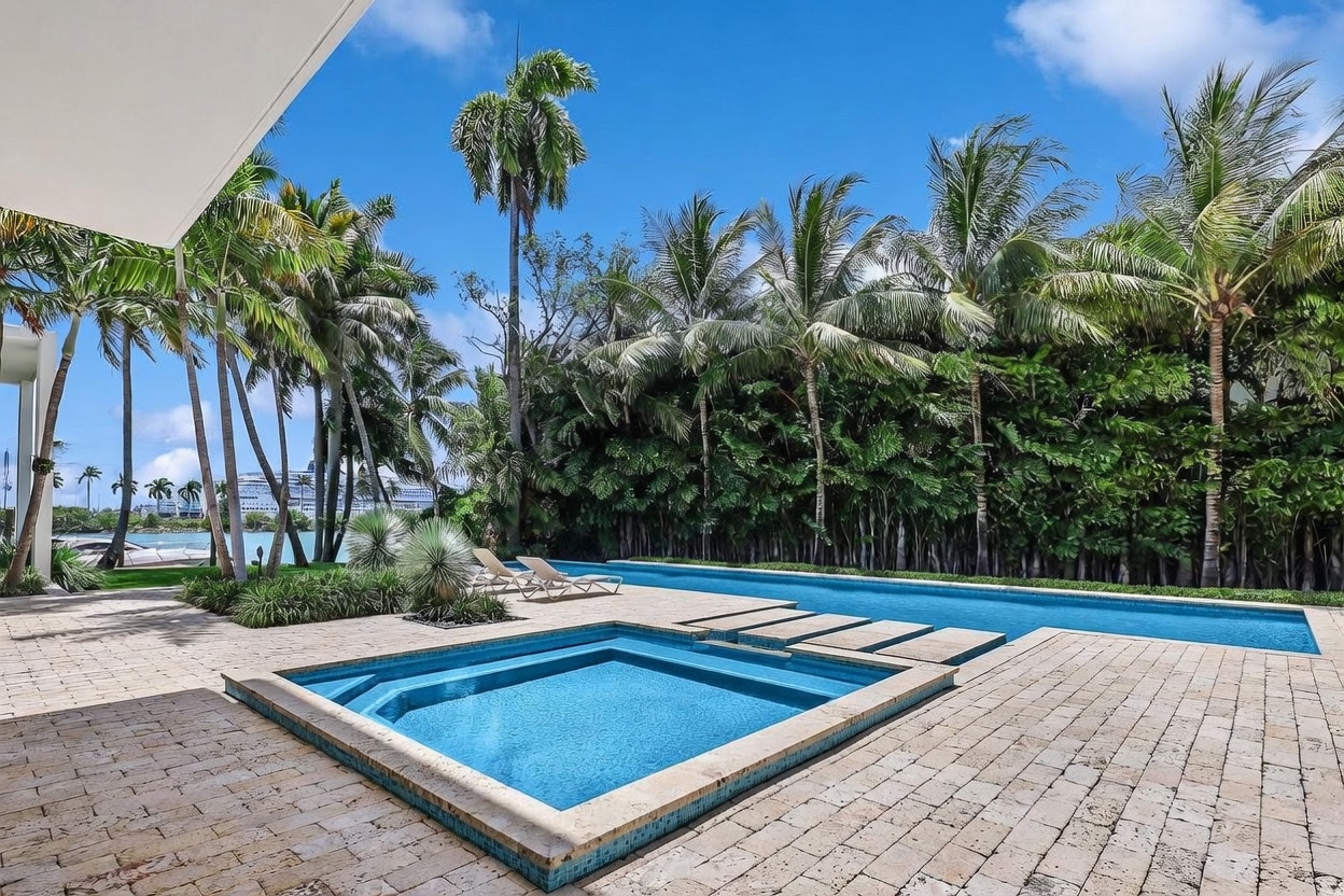 This luxurious outdoor space features a large, rectangular swimming pool paired with a square, integrated spa, both finished with vibrant blue tiles. The surrounding deck is paved with light-colored stone, and the area is framed by lush tropical palm trees and a dense green hedge. In the background, a glimpse of the waterfront and a large cruise ship adds a unique, high-end coastal atmosphere to the scene.