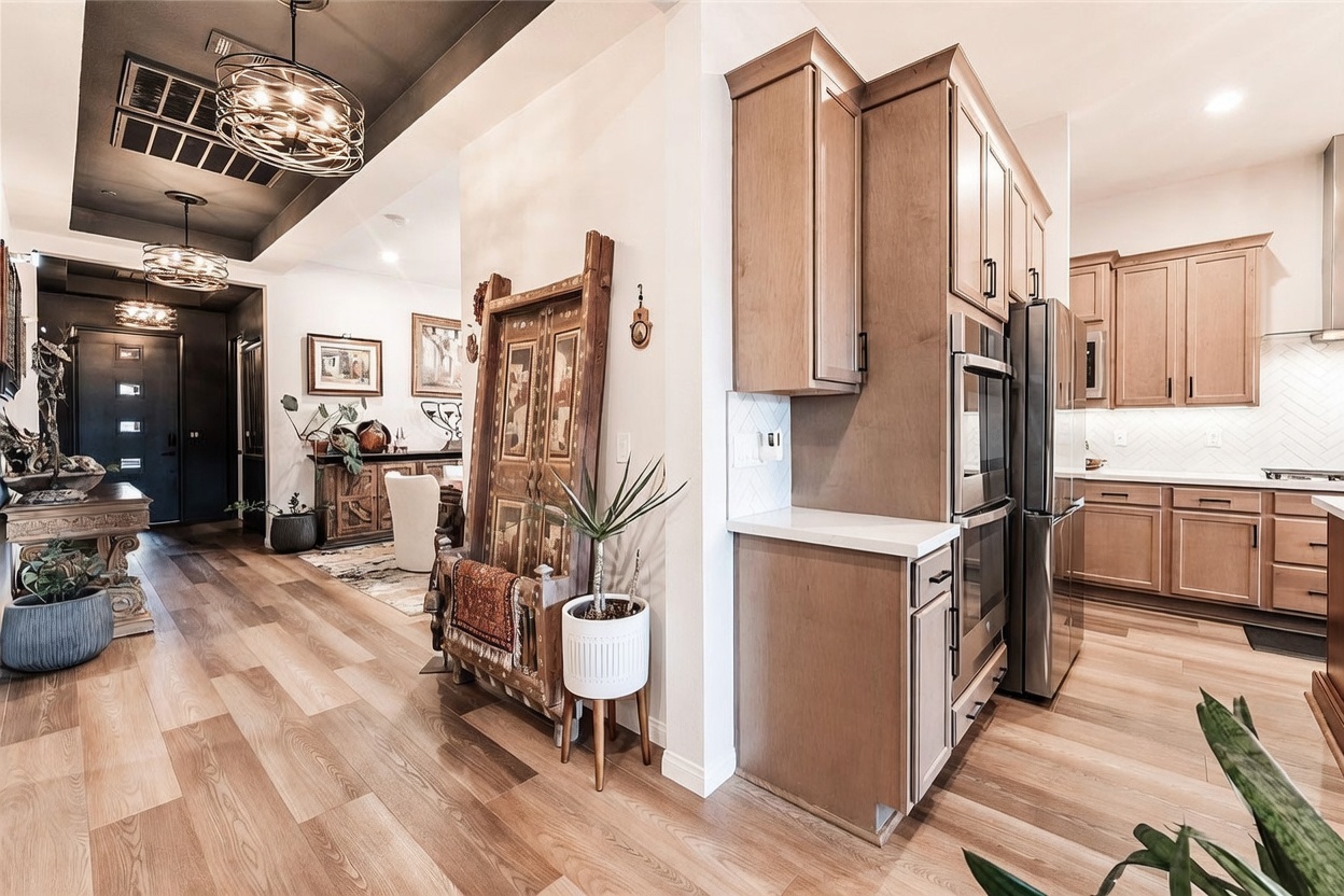 This interior view captures a spacious, open-concept kitchen and adjacent hallway area featuring light wood-toned flooring throughout. The kitchen is equipped with light-colored cabinetry, stainless steel appliances, and a clean, neutral aesthetic, while the foreground showcases a decorative wooden screen and a potted plant. The perspective looks from the kitchen toward a hallway and entryway, highlighting the home's cohesive design and warm, inviting atmosphere.