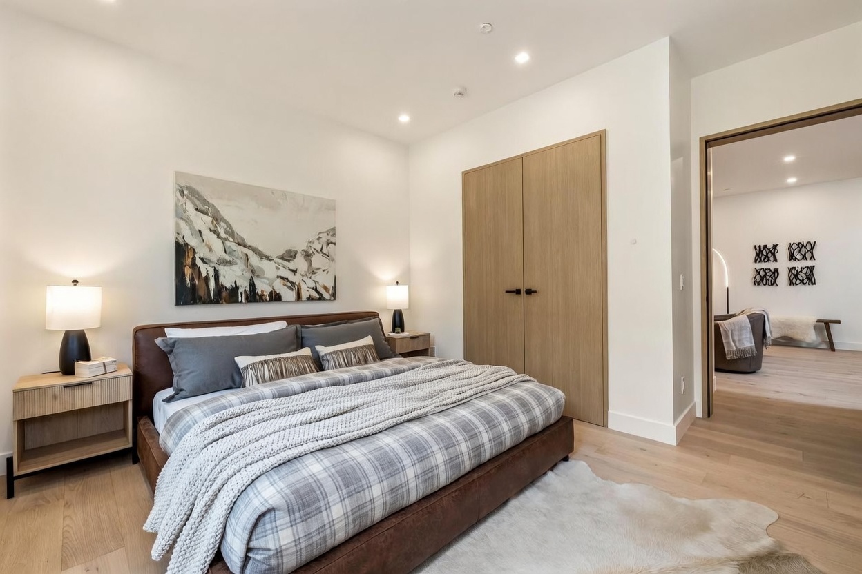 This contemporary bedroom features a low-profile leather bed frame topped with a plaid duvet and a textured throw blanket. The room is accented by light wood nightstands, matching table lamps, and a large mountain-themed landscape painting above the headboard. A light-toned area rug sits on the light wood flooring, while a doorway leads to an adjacent living space decorated with wall art.