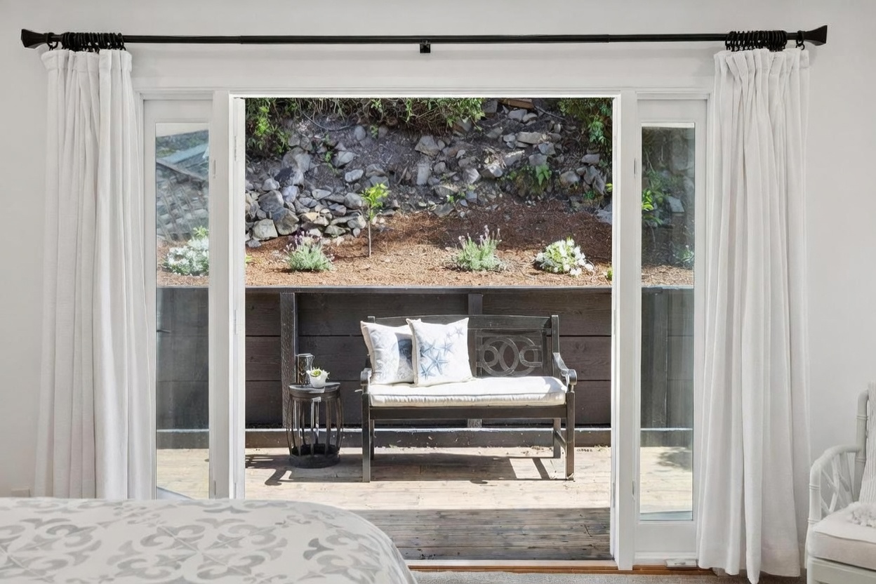 This view from a bedroom looks out through wide glass doors onto a private wooden deck featuring a bench and a small side table. The outdoor space is framed by a dark horizontal wood fence and a sloped, rocky hillside with natural landscaping. The interior is bright and neutral, with white curtains framing the doorway and a glimpse of a patterned bedspread in the foreground.