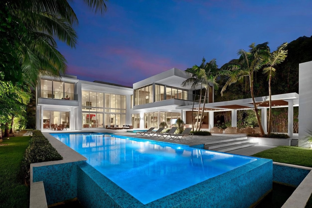 This stunning modern estate features a large, rectangular swimming pool as the centerpiece of its expansive rear terrace. The architecture is defined by clean lines, white structural elements, and extensive glass walls that create a seamless indoor-outdoor flow. Surrounded by lush tropical landscaping and illuminated by soft evening lighting, the property exudes a luxurious and serene atmosphere.