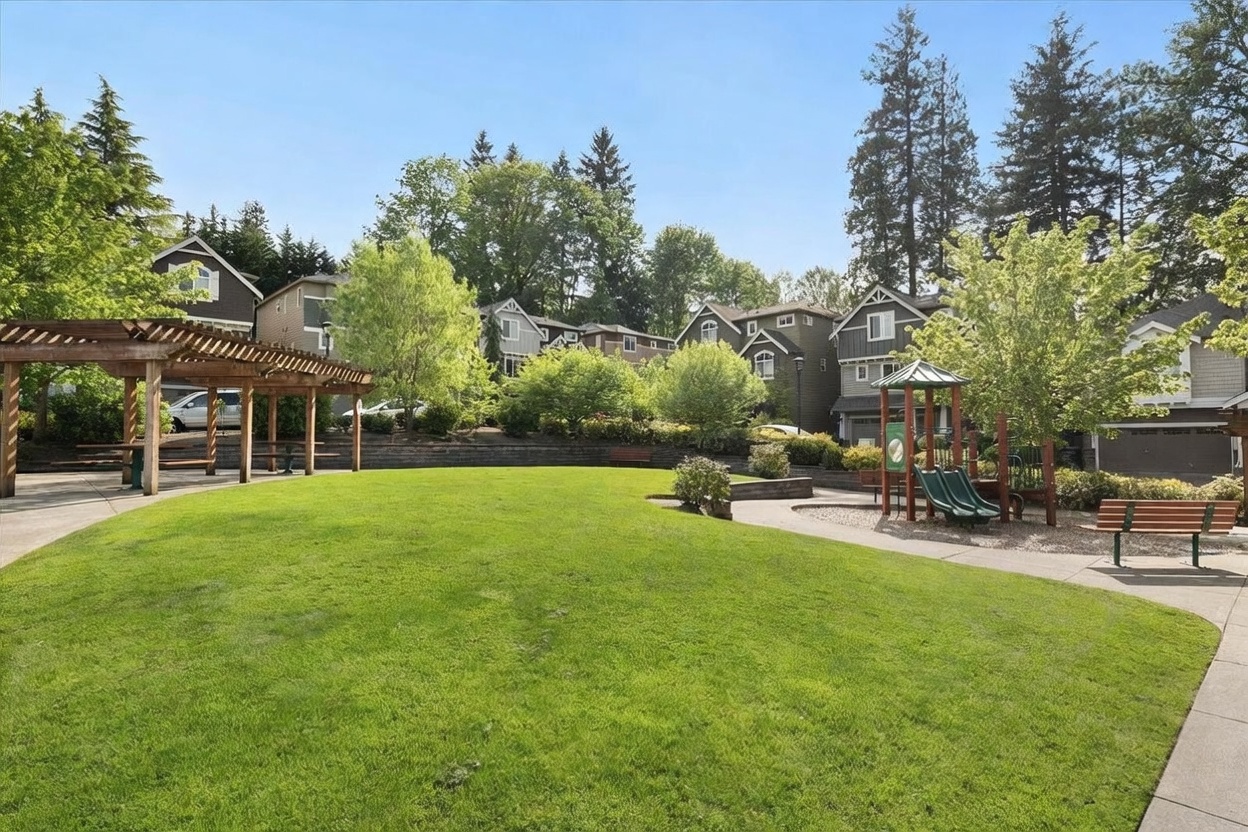 This image captures a well-maintained community park featuring a lush green lawn, a wooden pergola, and a children's playground area. The scene is framed by mature trees and residential townhomes in the background, creating a peaceful and inviting neighborhood atmosphere. The perspective is a wide-angle ground-level shot that emphasizes the open space and recreational amenities available to residents.