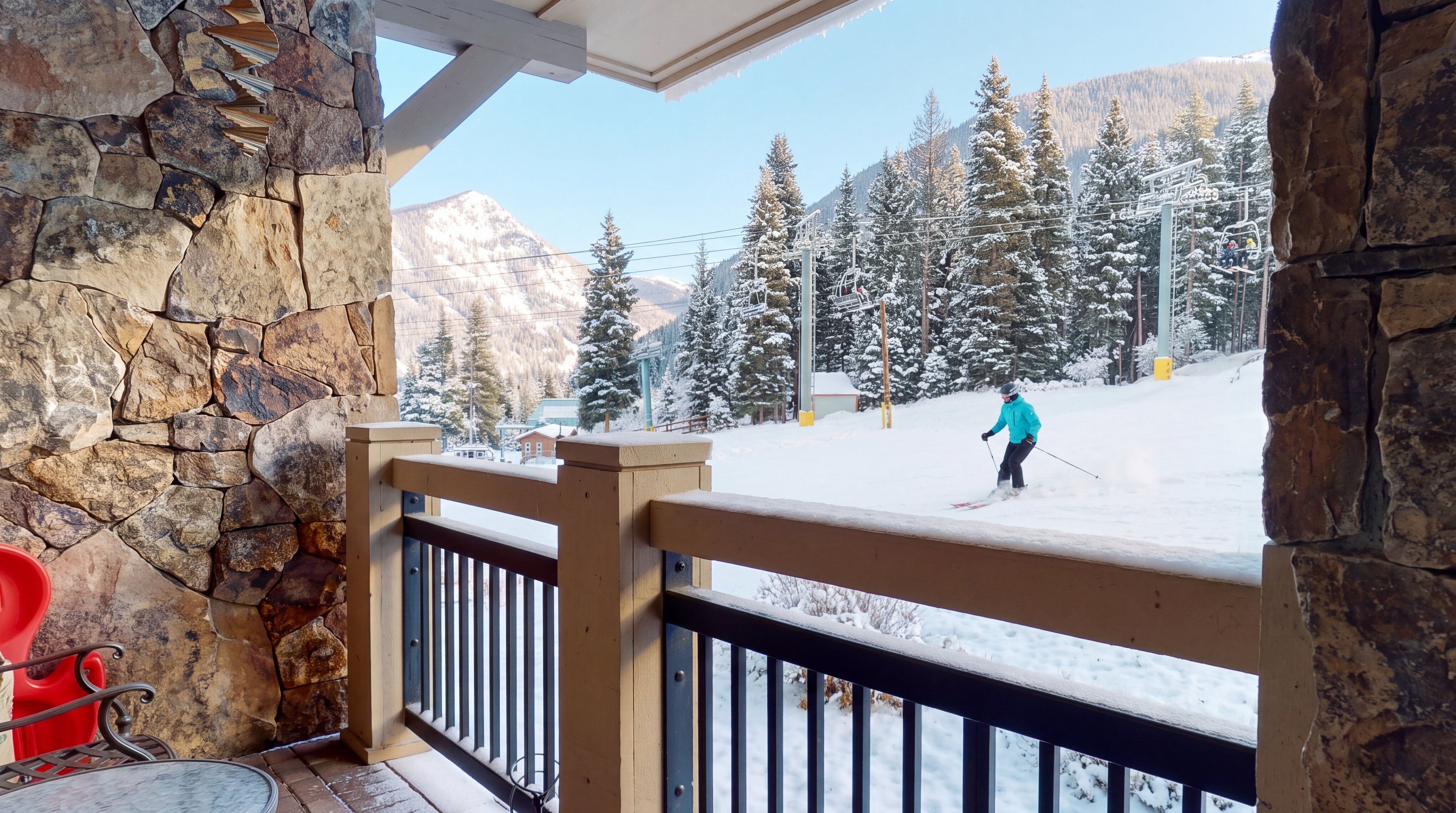 This covered balcony offers a scenic view of a mountain landscape, complete with ski lift infrastructure visible in the distance. The space features a stone-clad wall, a brick-paved floor, and a metal railing, with a small bistro table and a red plastic chair providing a casual seating area. The perspective looks outward from the sheltered deck toward the grassy slope and forested mountains under a clear blue sky.