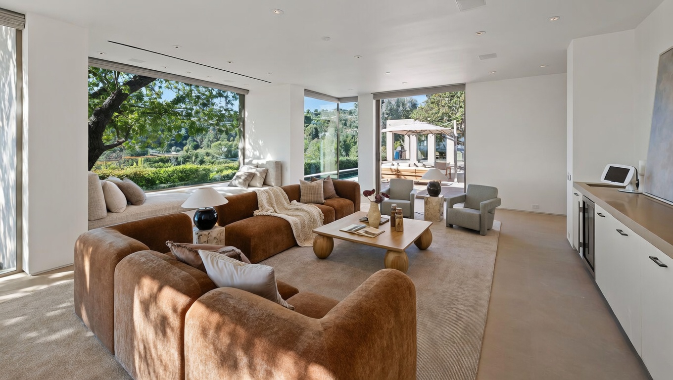 This spacious, modern living room features a large, plush brown sectional sofa arranged around a minimalist wooden coffee table. Expansive floor-to-ceiling windows flood the room with natural light and offer serene views of the lush outdoor landscape. The neutral color palette and clean lines create an airy, sophisticated atmosphere perfect for relaxation and entertaining.