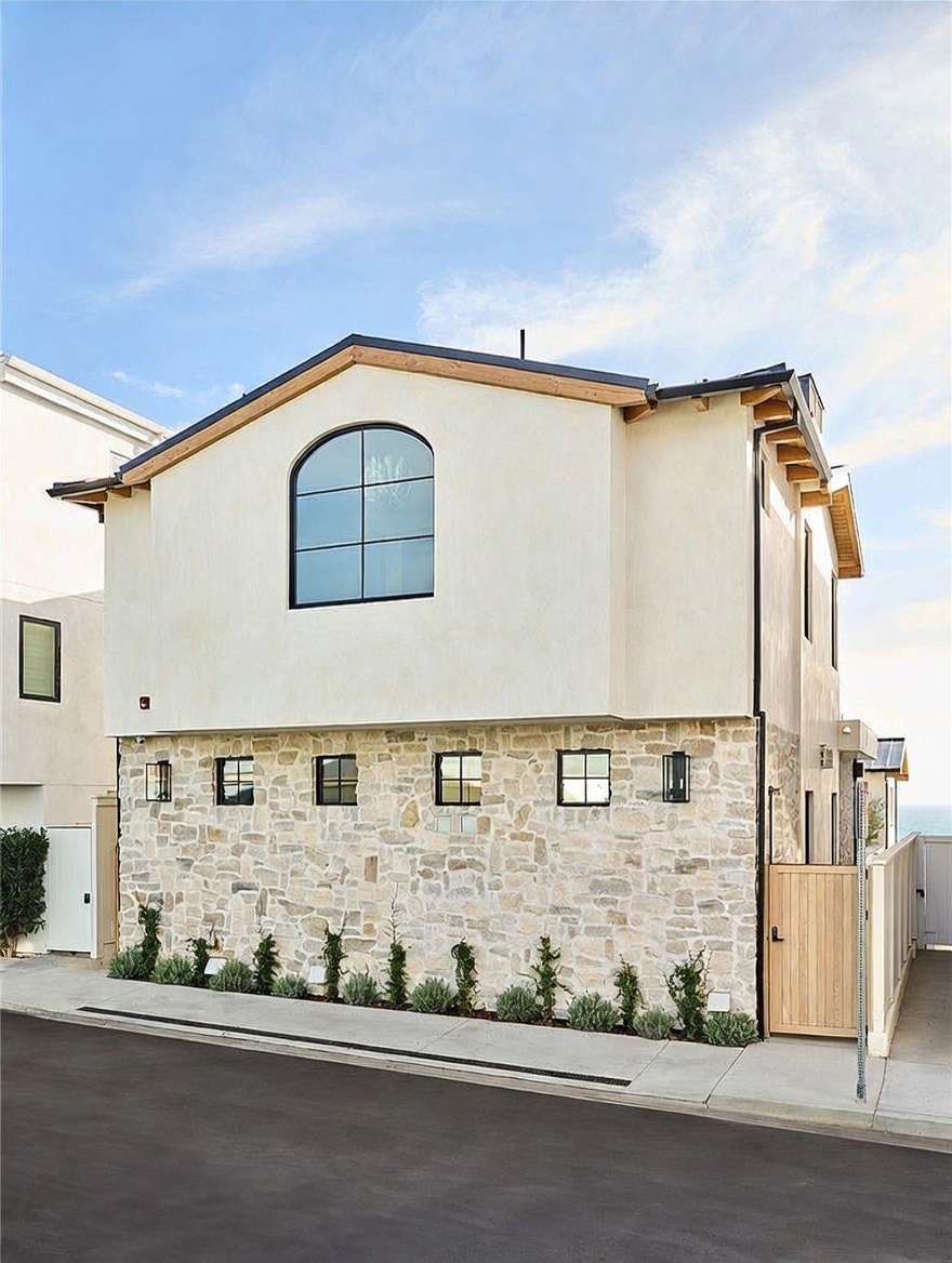 This side view of a modern coastal home showcases a striking contrast between smooth, light-colored stucco on the upper level and a textured, natural stone facade on the lower level. A prominent arched window serves as a focal point on the upper floor, while a series of rectangular windows lines the stone base. The property is framed by a clean concrete sidewalk and a neatly landscaped strip of low-lying shrubs, leading to a wooden gate on the right.