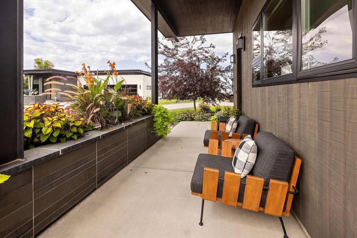 This modern covered porch features a sleek concrete floor and a long, built-in planter box filled with vibrant flowers and greenery. Two contemporary wooden chairs with dark cushions and plaid pillows are arranged to overlook the front yard, creating a cozy and inviting outdoor seating area. The exterior wall is clad in vertical wood-look siding, and the space is framed by a dark metal support column, emphasizing a clean, architectural aesthetic.