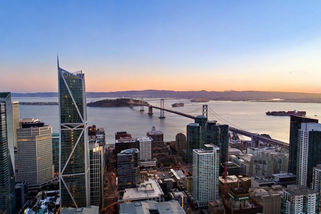 This high-altitude aerial view captures a stunning cityscape of San Francisco at dusk, featuring the iconic Bay Bridge stretching across the water. The foreground is dominated by modern high-rise architecture, including a prominent glass-clad skyscraper with a distinct geometric design. The soft, warm glow of the setting sun illuminates the horizon, creating a dramatic and sophisticated urban atmosphere.