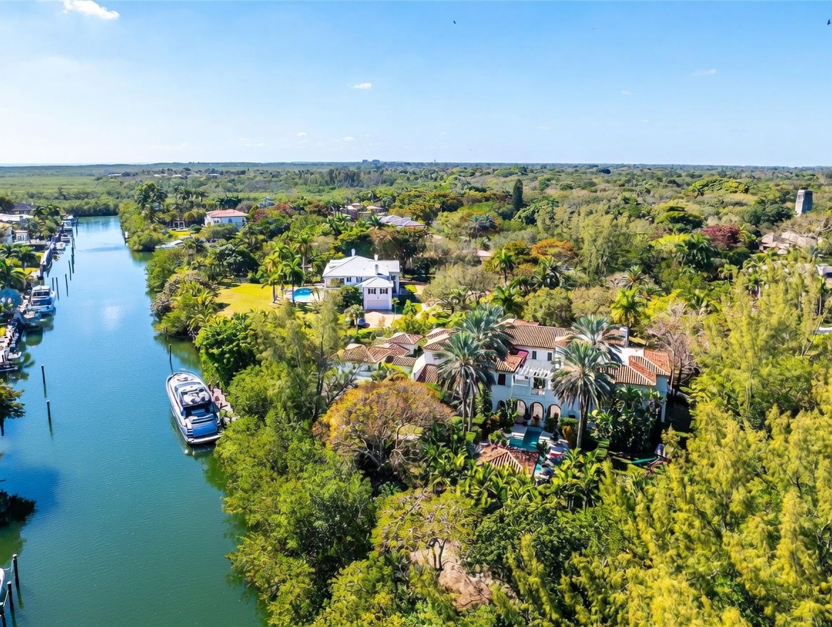 This high-angle aerial perspective captures a sprawling Mediterranean-style estate nestled within a lush, tropical landscape. The property features a multi-level residence with a terracotta tile roof, a private swimming pool, and direct access to a tranquil waterway where a yacht is docked. The surrounding environment is dense with mature trees and greenery, emphasizing a secluded and luxurious waterfront lifestyle.