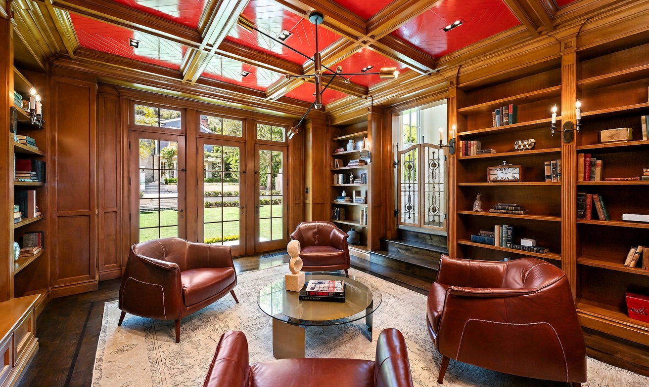 This sophisticated study features rich wood-paneled walls lined with built-in bookshelves and a striking, high-gloss red coffered ceiling. Three leather armchairs are arranged around a central glass-topped coffee table, creating an inviting conversation area. Large glass-paned doors provide a view of the outdoor greenery, while a modern geometric chandelier hangs as a focal point above the room.
