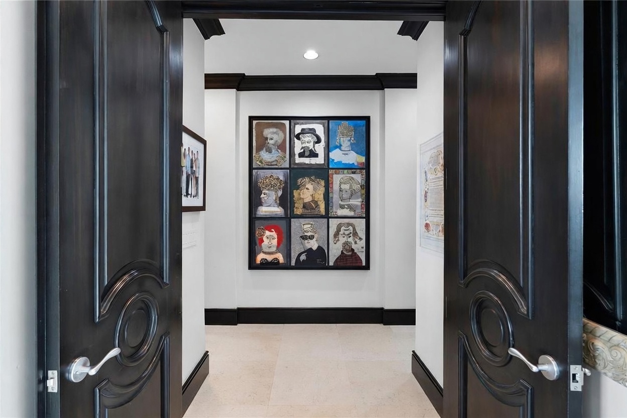 This interior view captures a formal entryway or hallway framed by two large, dark-stained wooden doors with ornate carved detailing and silver-toned hardware. The perspective looks through the open doorway toward a white wall featuring a large, multi-paneled piece of contemporary art depicting various stylized portraits. The space is finished with light-colored stone flooring and dark crown molding, creating a high-contrast, sophisticated aesthetic.