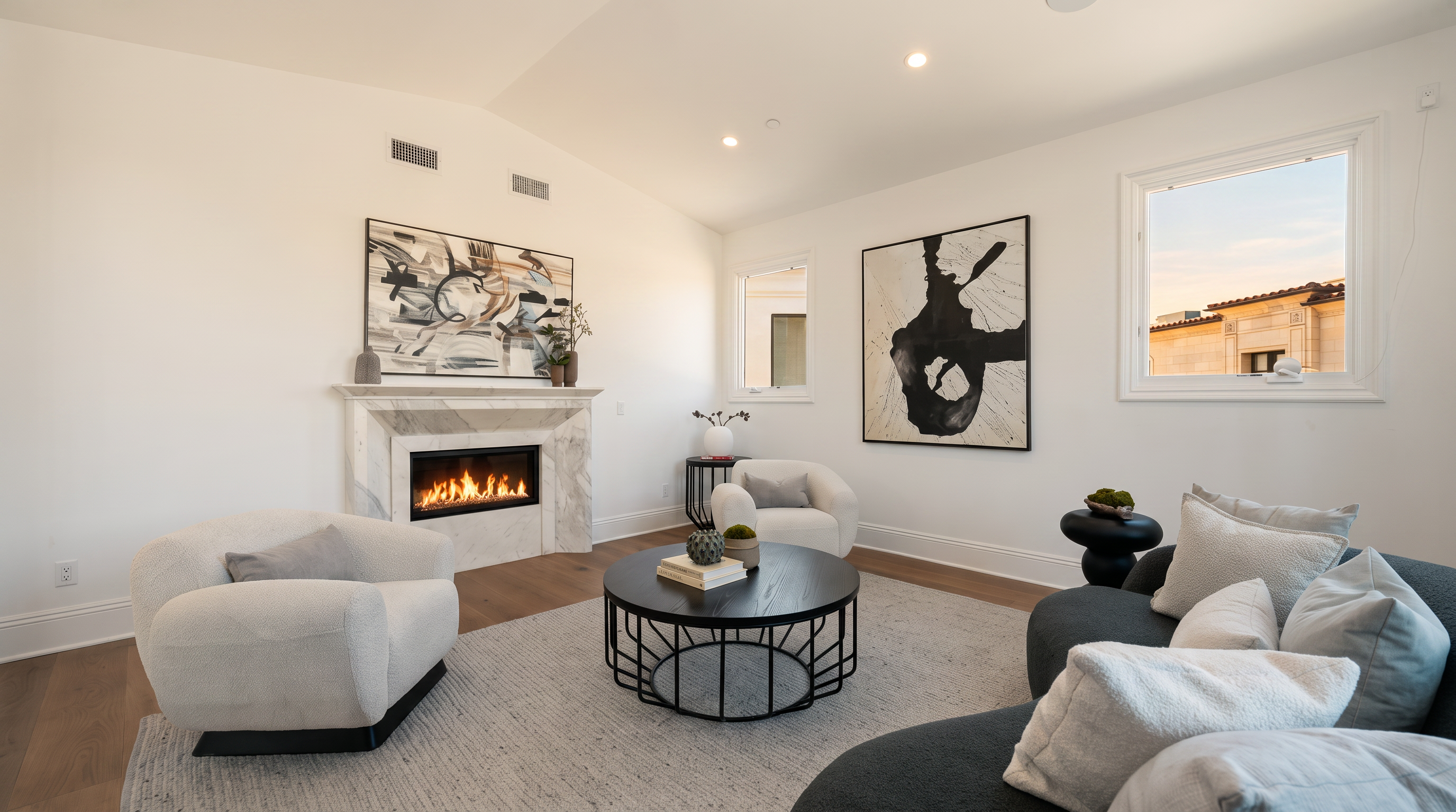 This bright and airy living room features a contemporary design with a neutral color palette and light wood flooring. A central fireplace with a stone surround and mantel serves as a focal point, complemented by abstract artwork on the walls. The space is furnished with a curved sofa, a round coffee table, and two plush armchairs, creating a comfortable and inviting seating area.