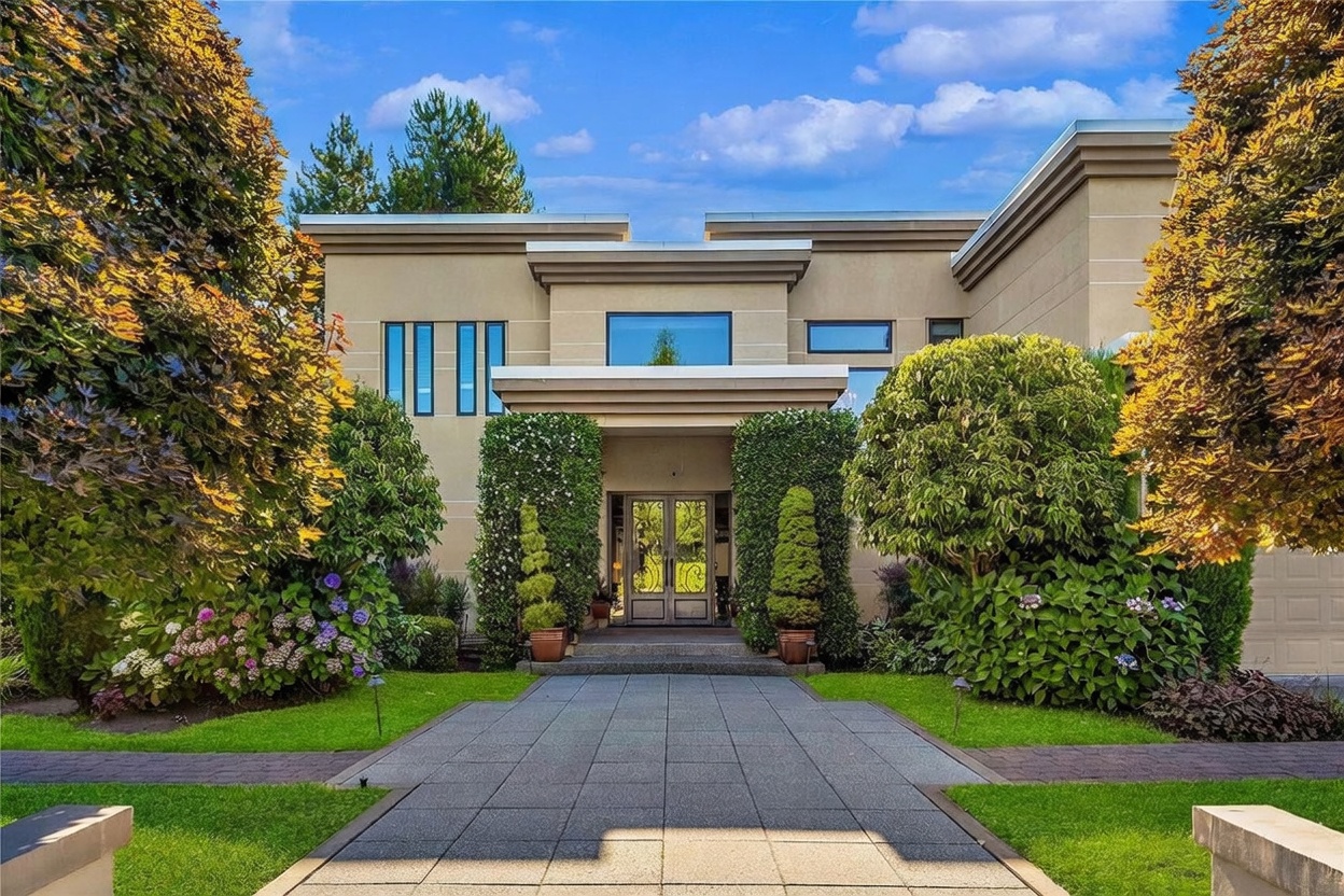 This modern, multi-level residence features a symmetrical facade with clean lines and a neutral stucco finish. A paved walkway leads directly to a grand double-door entrance framed by manicured hedges and potted topiary, while mature trees and lush landscaping flank the front yard. The composition is captured from a low-angle, eye-level perspective, emphasizing the home's architectural presence and curb appeal.