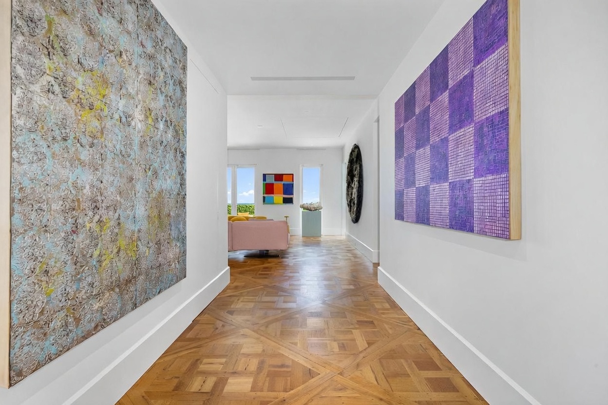 This elegant hallway features intricate, light-toned wood parquet flooring and crisp white walls adorned with large, vibrant abstract art pieces. The perspective draws the eye through the gallery-like corridor toward a bright living area at the end, which is illuminated by natural light from large windows. The space exudes a sophisticated, gallery-inspired aesthetic, emphasizing clean lines and a curated atmosphere.